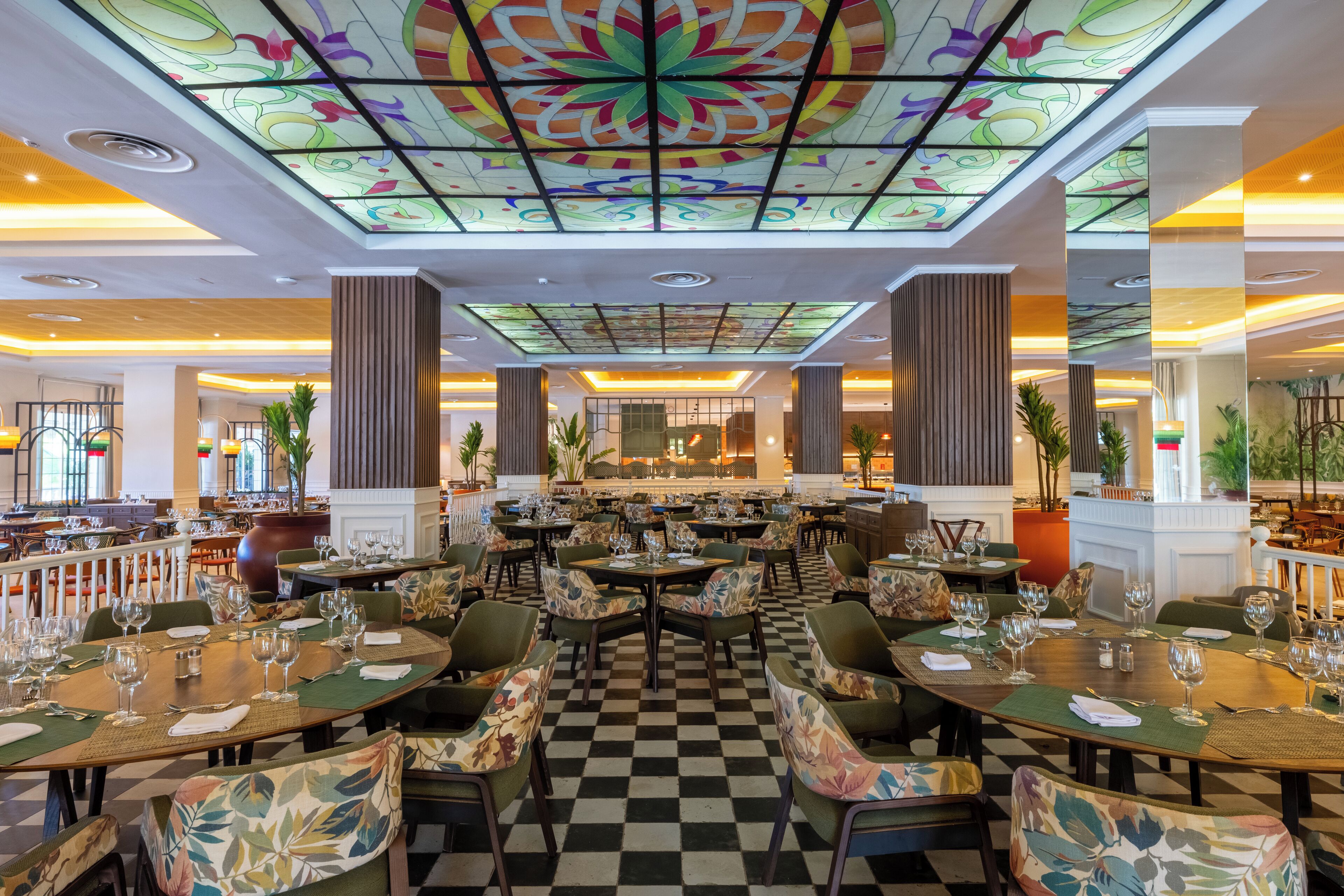 A restaurant with checkered flooring, floral chairs, and a stained glass ceiling.