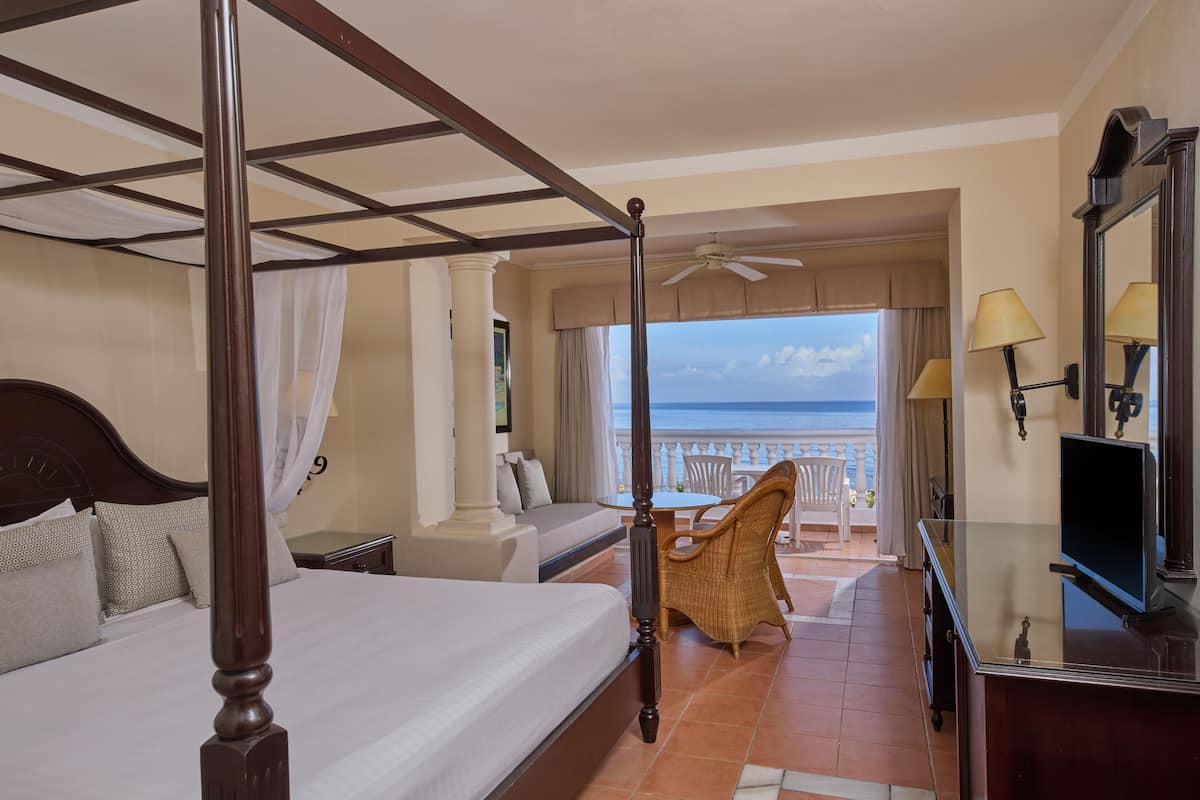 A four-poster bed, a sofa, a dining table with chairs, and a TV in a hotel room with a balcony overlooking the sea.
