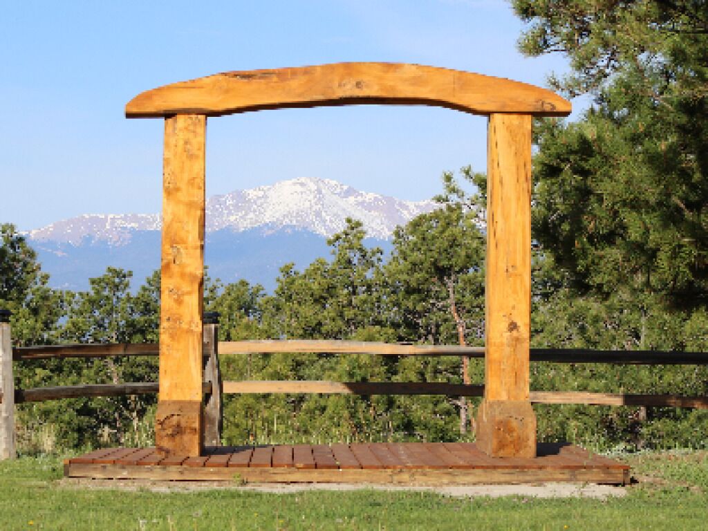 A wooden arch structure with a wooden deck in front, set against a backdrop of trees and mountains.