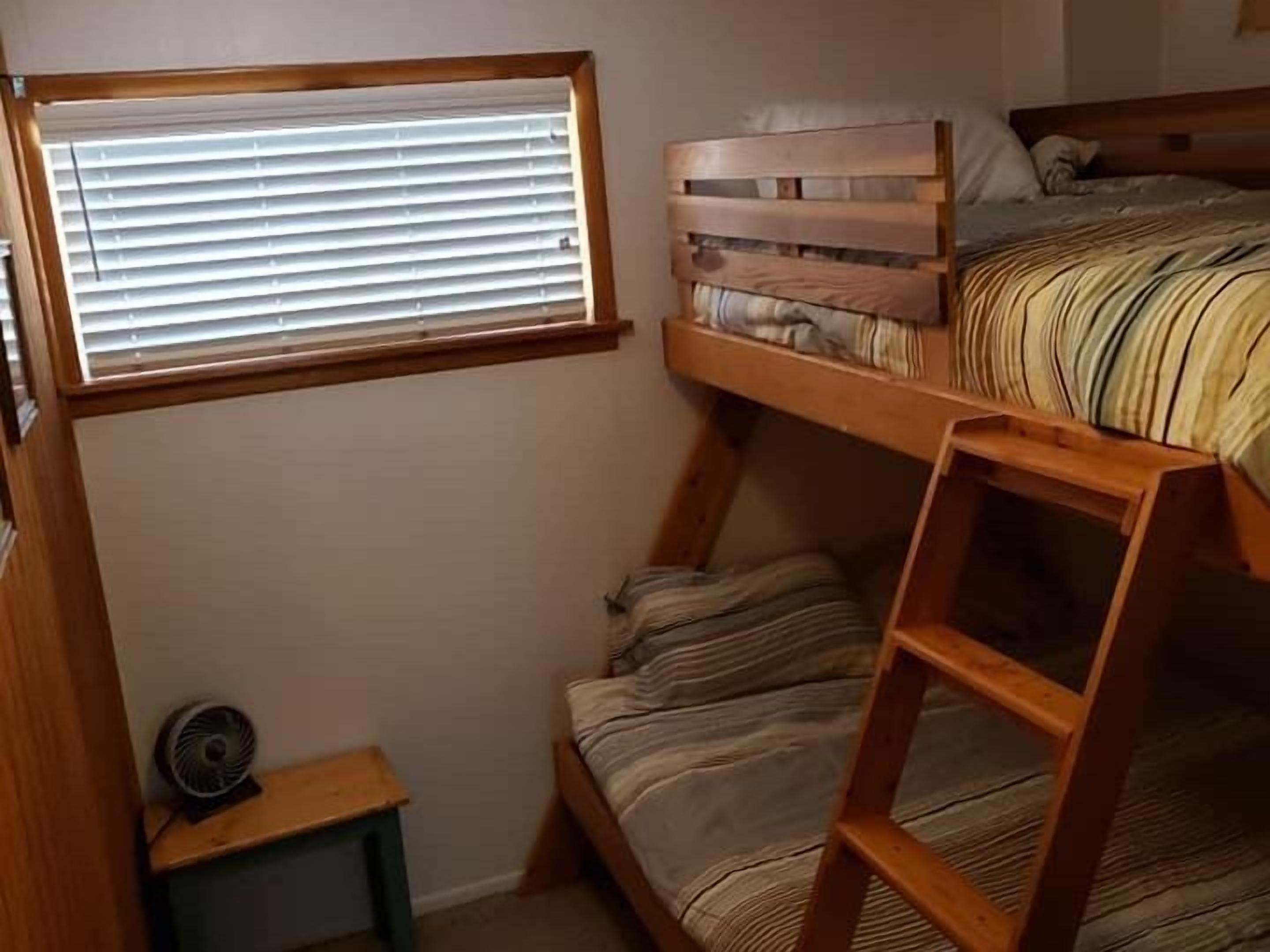 A bunk bed with a wooden ladder, a small table with a fan, and a window with blinds.