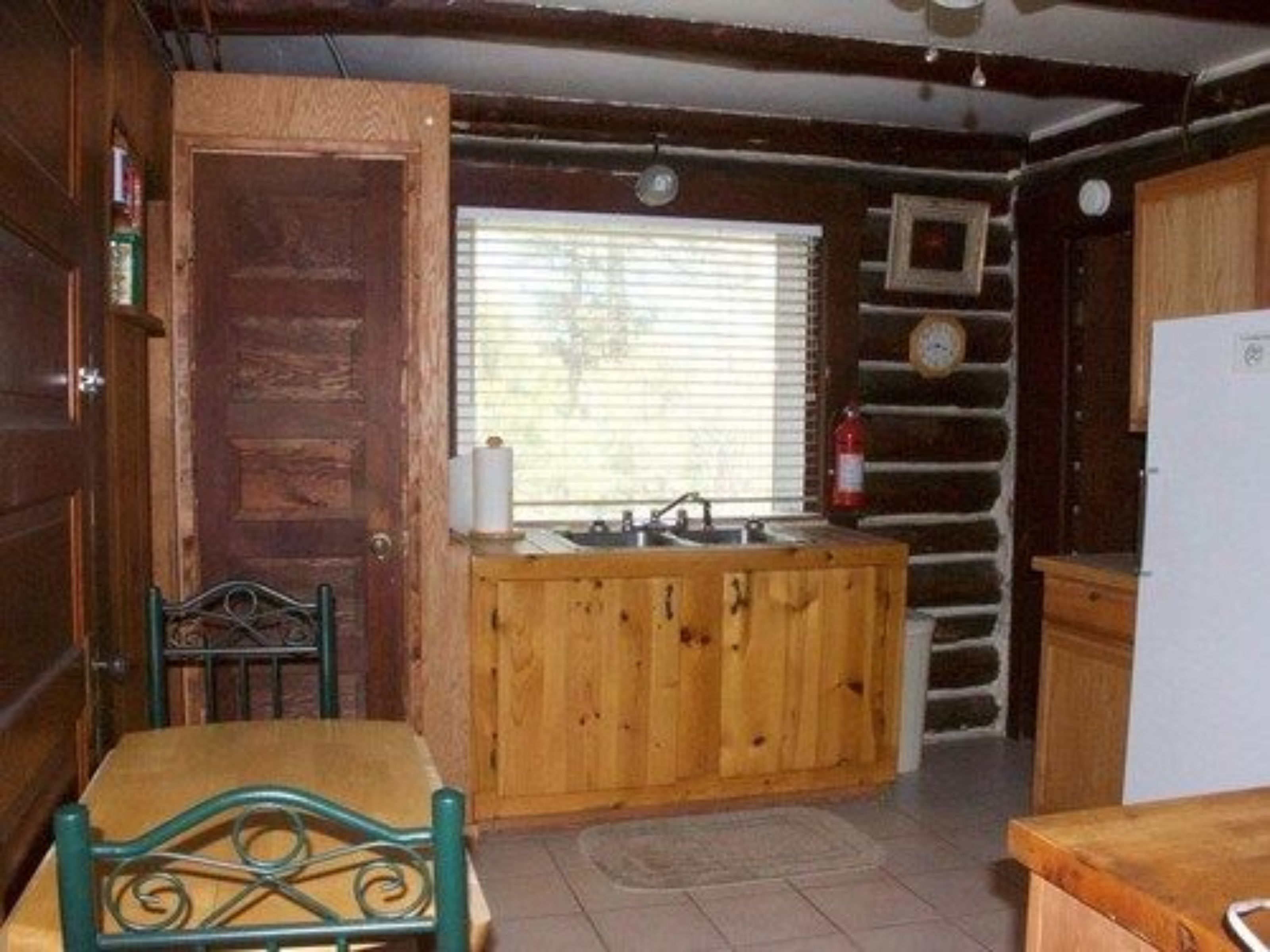 A rustic cabin interior with a wooden dining table, a bench, a kitchen area with a sink and a refrigerator, and a window with blinds.