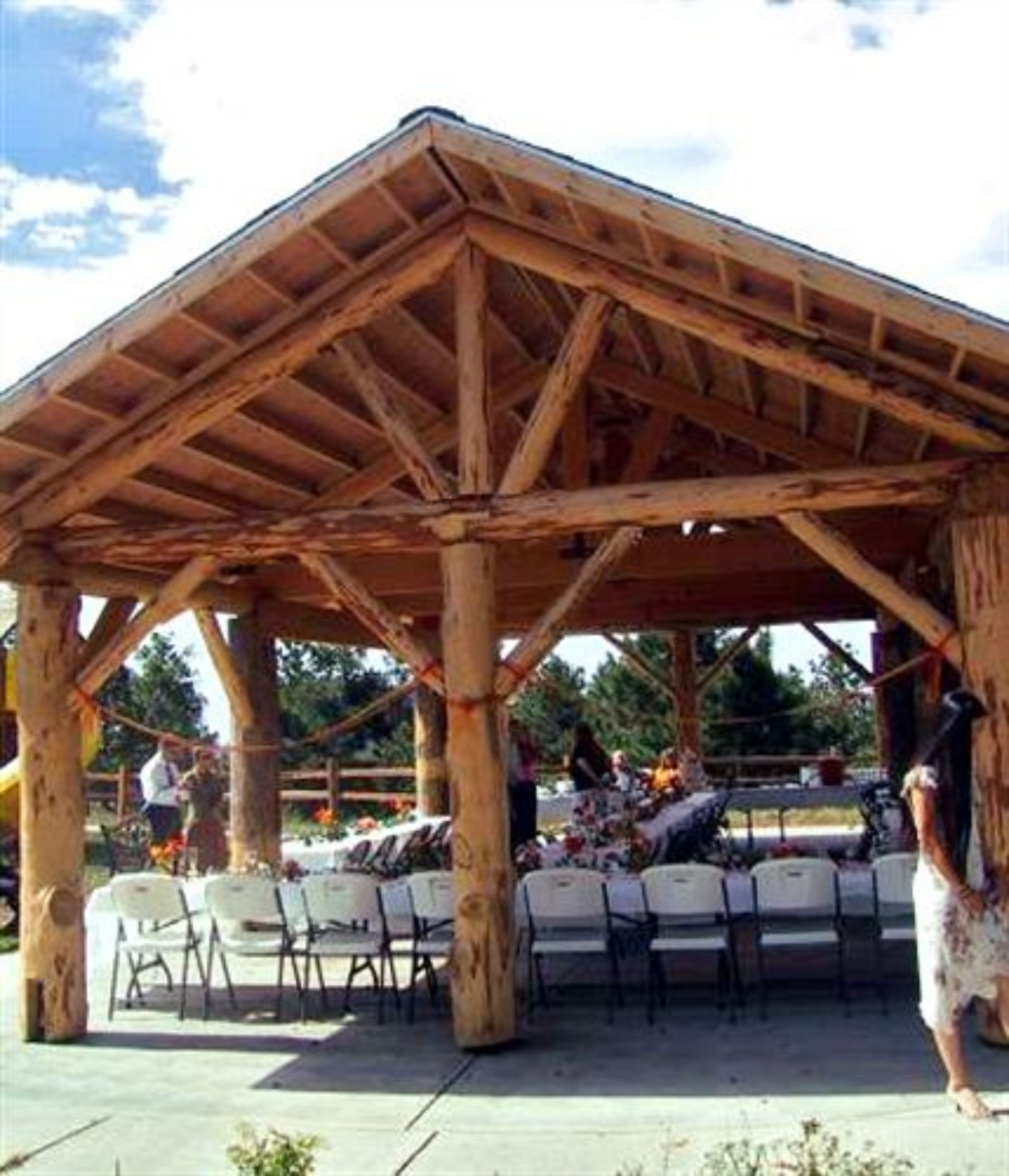 Un pabellón de madera con estructura de techo sencilla, rodeado de sillas y mesas de plástico blanco dispuestas para un evento.