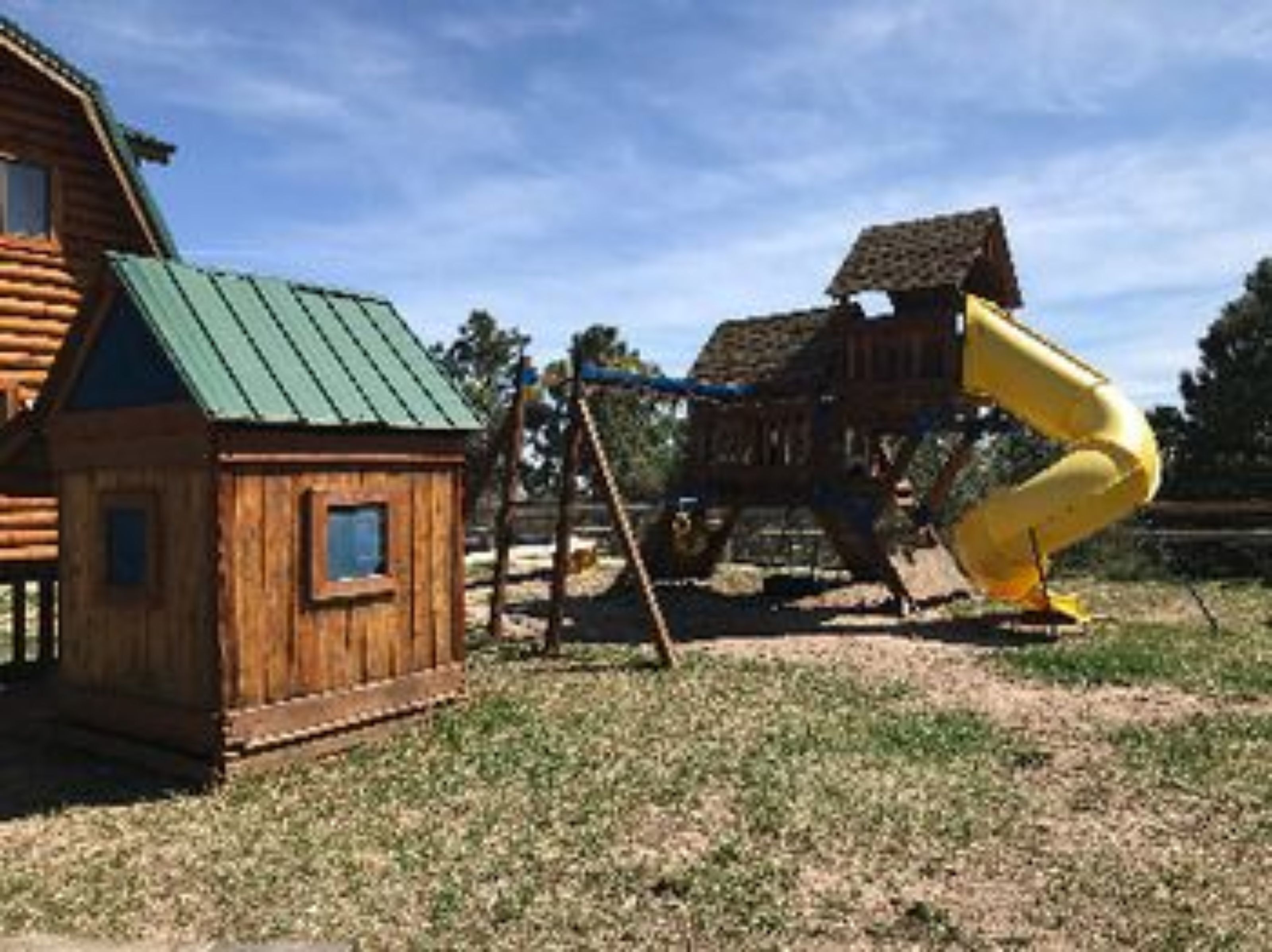 Une cabane en bois avec un toit vert, une petite fenêtre et une structure de jeu en bois dotée d’un toboggan jaune.