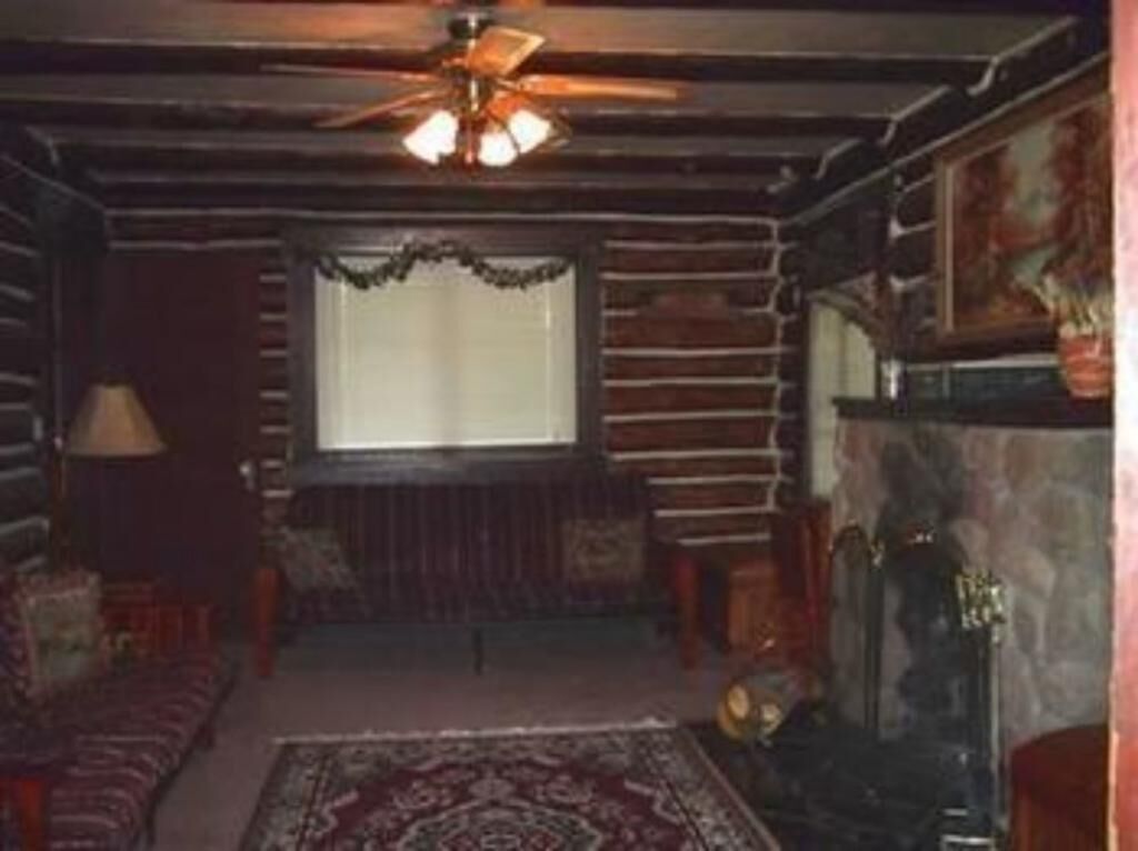 A log cabin interior with a fireplace, a ceiling fan, and a window with curtains.