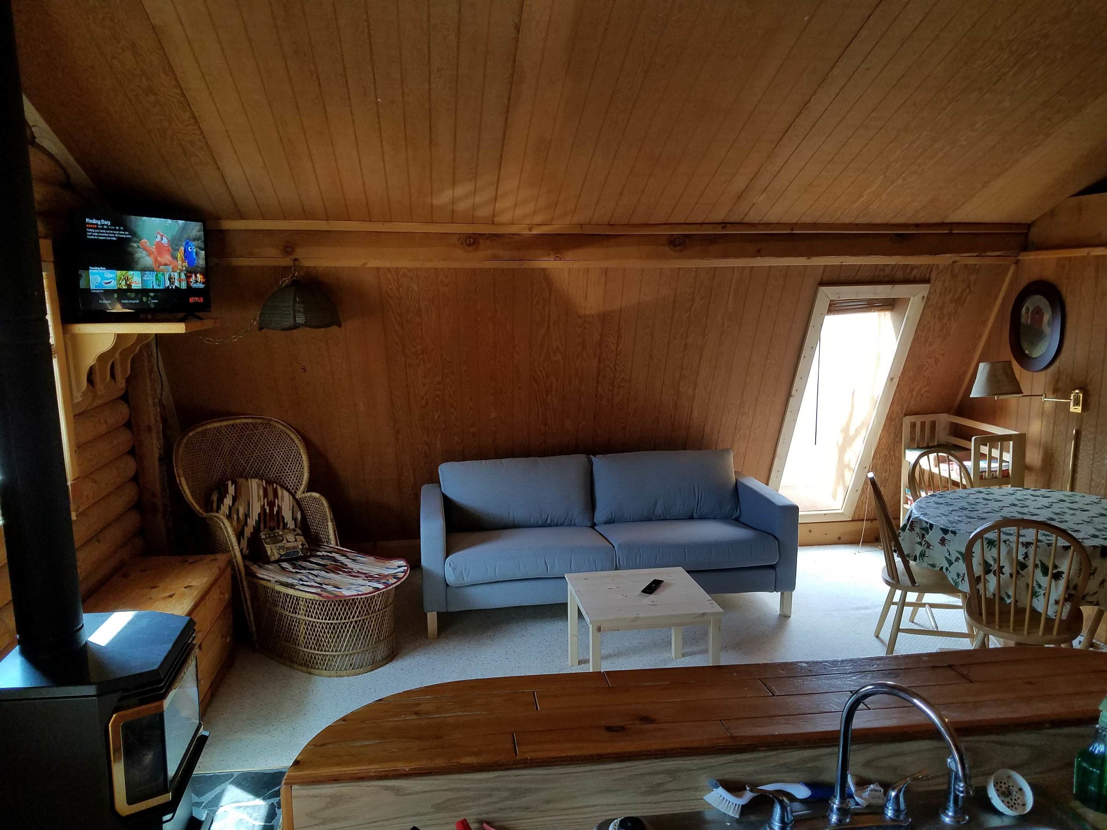 A cozy log cabin interior with a blue sofa, a small table, a wicker chair, a wooden dining table with chairs, a TV mounted on a wooden beam, and a window with a view of the outdoors.