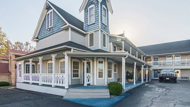 A large, two-story house with a steeply pitched roof, multiple windows, and a porch with white railings. - Motel 6 Paris, TX (Paris)