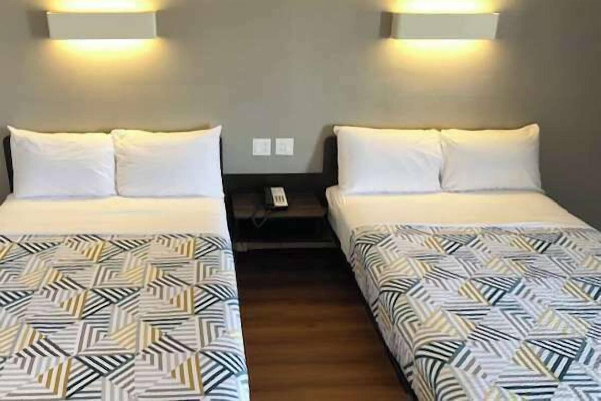 A hotel room with two beds, each with a patterned bedspread and white pillows. There are wall-mounted light fixtures above each bed.