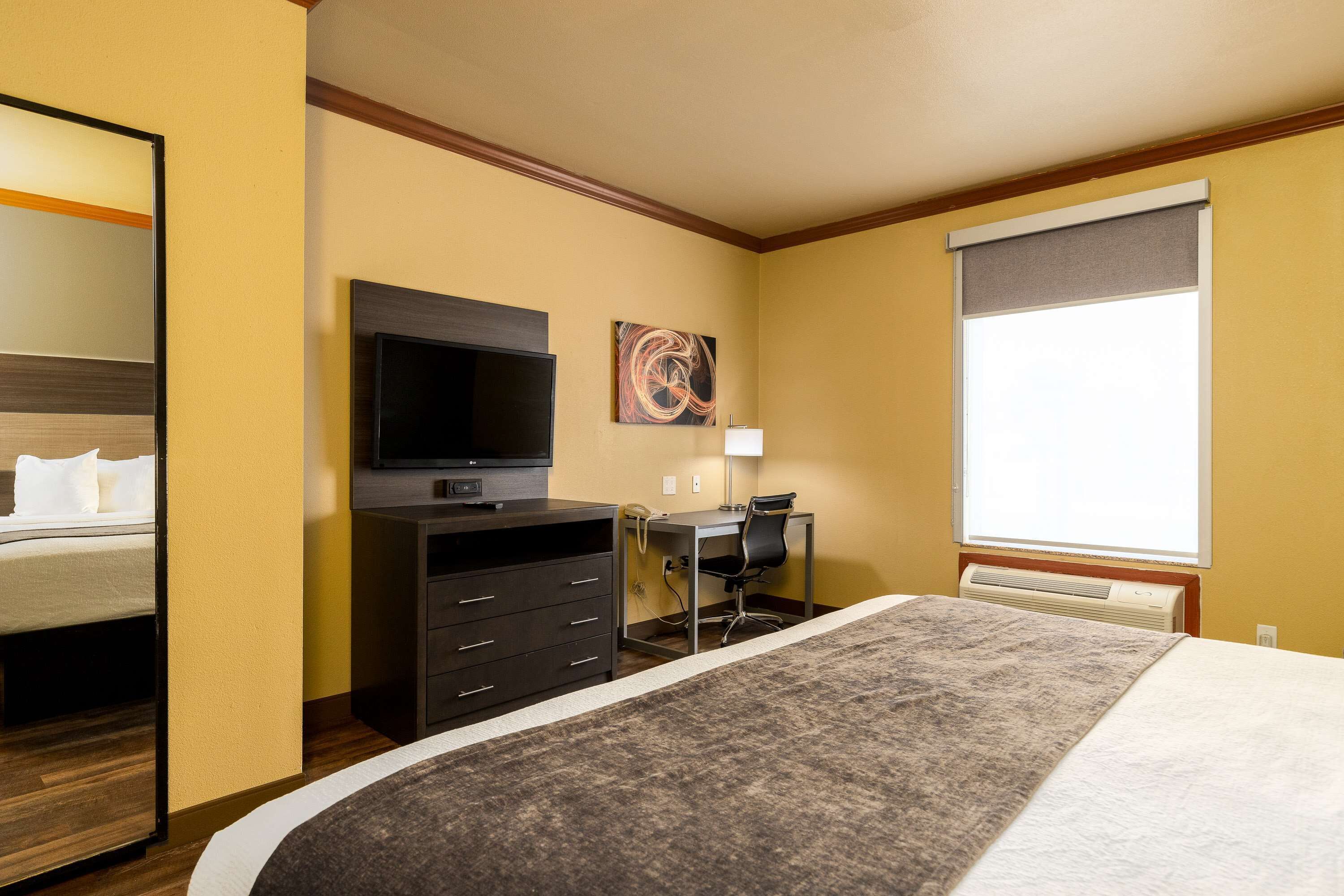 A hotel room with a bed, a desk with a chair, a television, and a window with blinds.