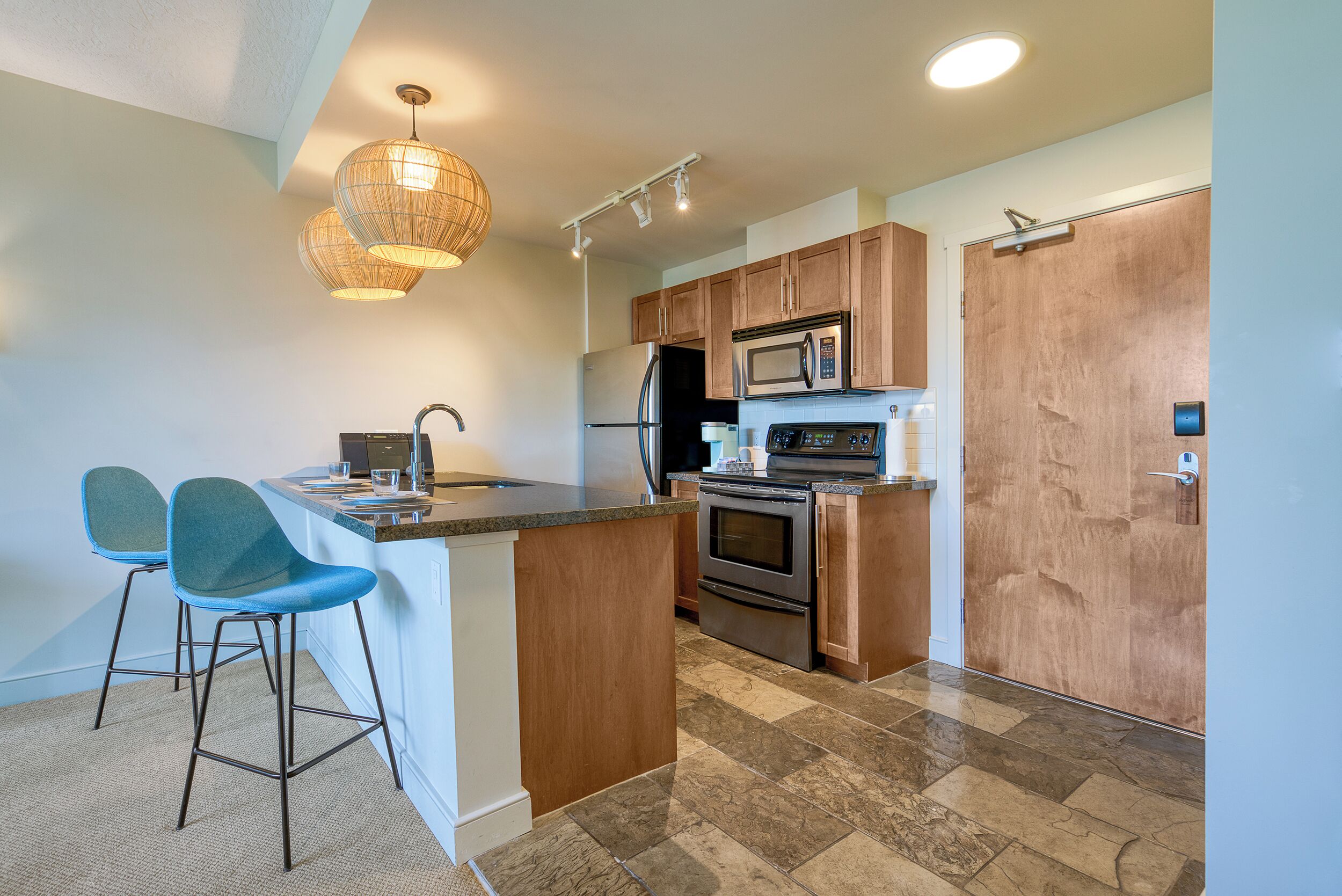 Una cocina moderna con una isla central, dos taburetes de bar, armarios de madera, electrodomésticos de acero inoxidable y una lámpara colgante.