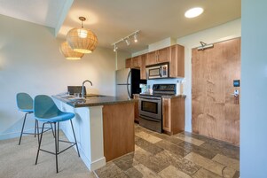 Una cocina moderna con una isla central, dos taburetes de bar, armarios de madera, electrodomésticos de acero inoxidable y una lámpara colgante.