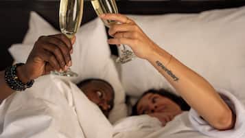 Two people toasting with champagne glasses in bed, with a visible tattoo on one arm.