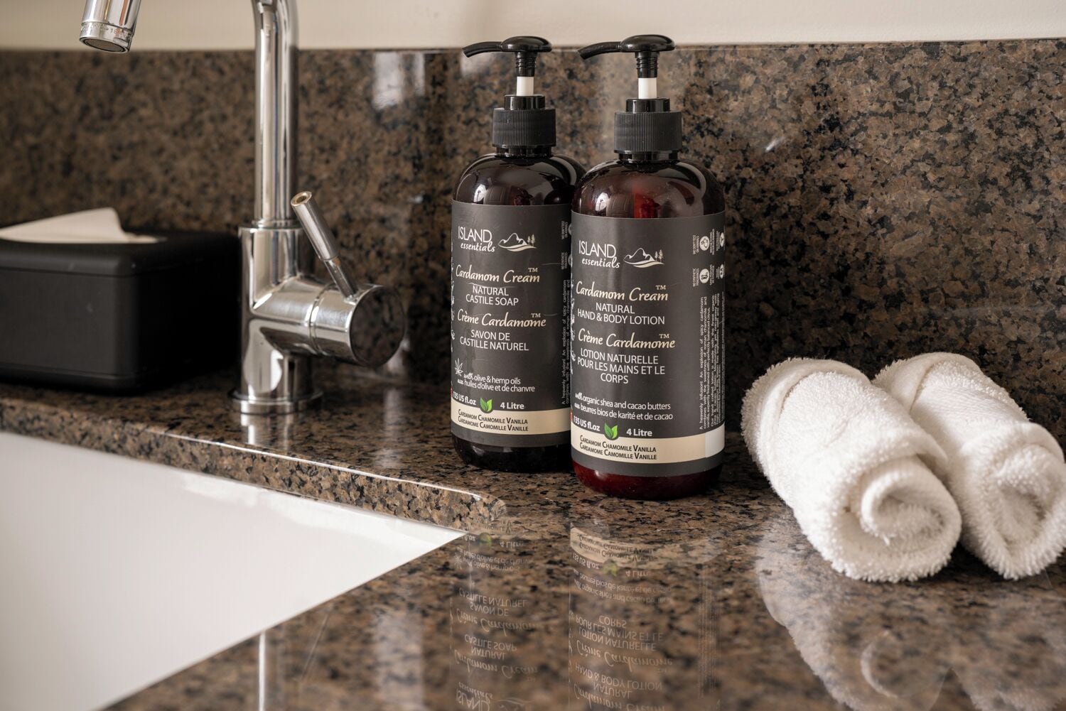 Dos botellas de jabón y loción Cardamom Cream de Island Essentials sobre una encimera de granito, junto a un lavabo y una toalla blanca enrollada.