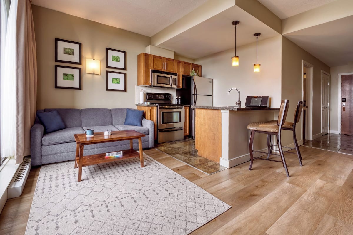 Una sala de estar moderna con un sofá gris, una mesa de centro de madera y un tapete estampado. Se ve una cocina con armarios de madera, un microondas y un refrigerador al fondo.