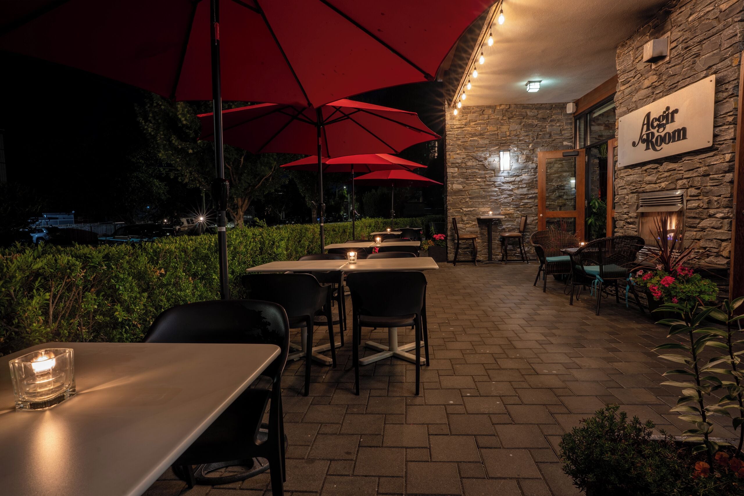 Een terras buiten met tafels en stoelen onder rode parasols, stenen muren en een bordje met de tekst 