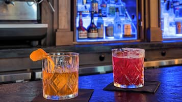 Dos vasos de bebidas sobre una barra, con estantes al fondo llenos de botellas.