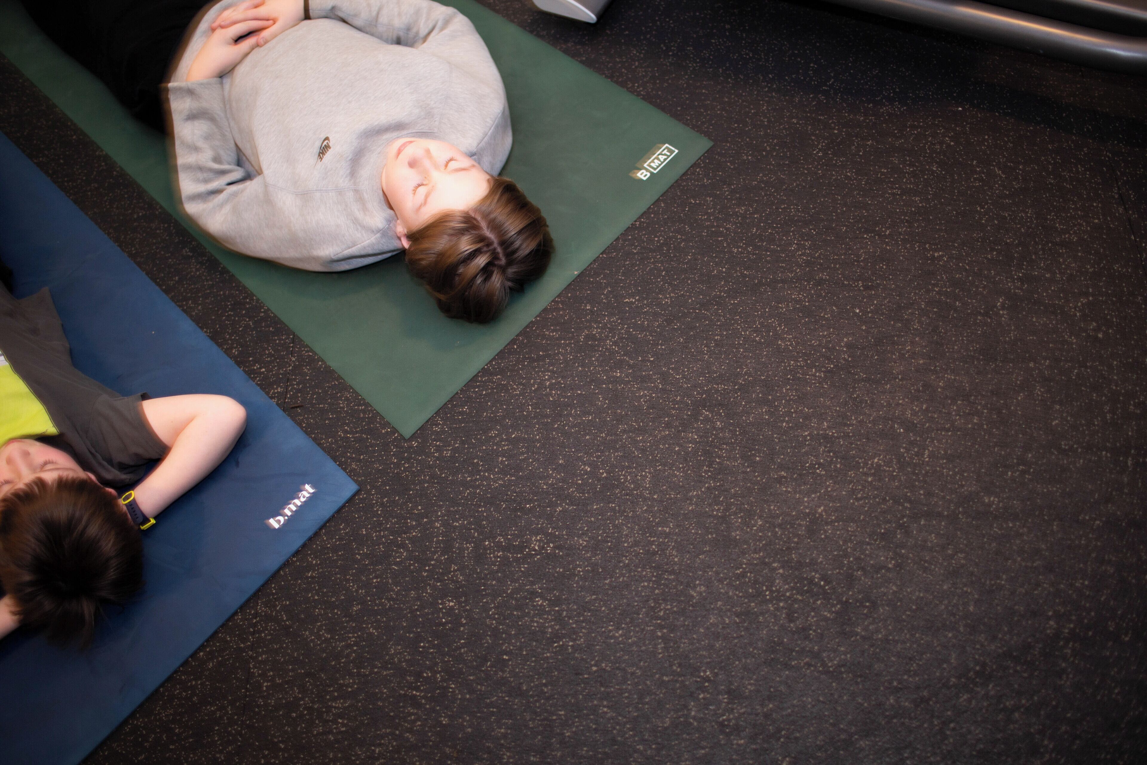 A person lying on a green exercise mat in a room with a speckled floor.