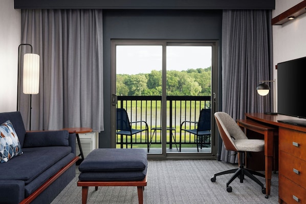 A hotel room with a sofa, a desk, a chair, and a large window with a view of a green landscape.