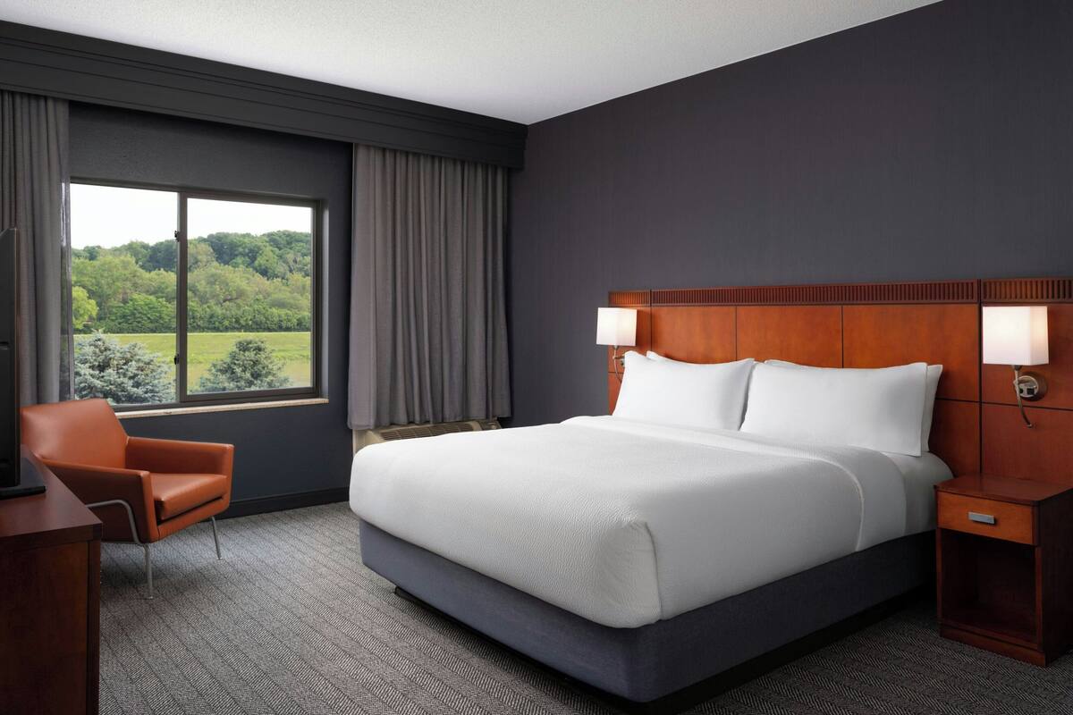 Ein Hotelzimmer mit einem großen Bett, einem Schreibtisch, einem Stuhl und Blick auf Grün durch das Fenster.