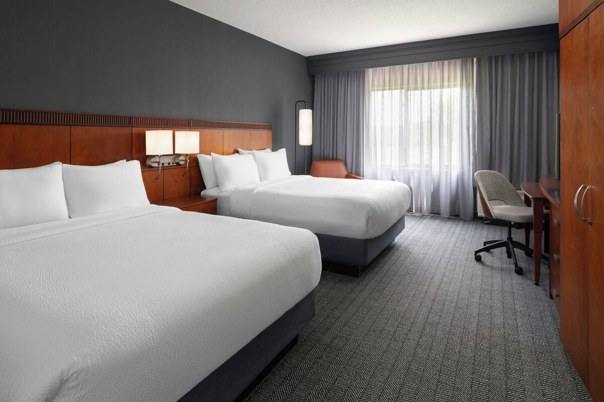 Ein Hotelzimmer mit zwei Betten, einem Schreibtisch, einem Stuhl, einem Kleiderschrank und einem großen Fenster mit Vorhängen.