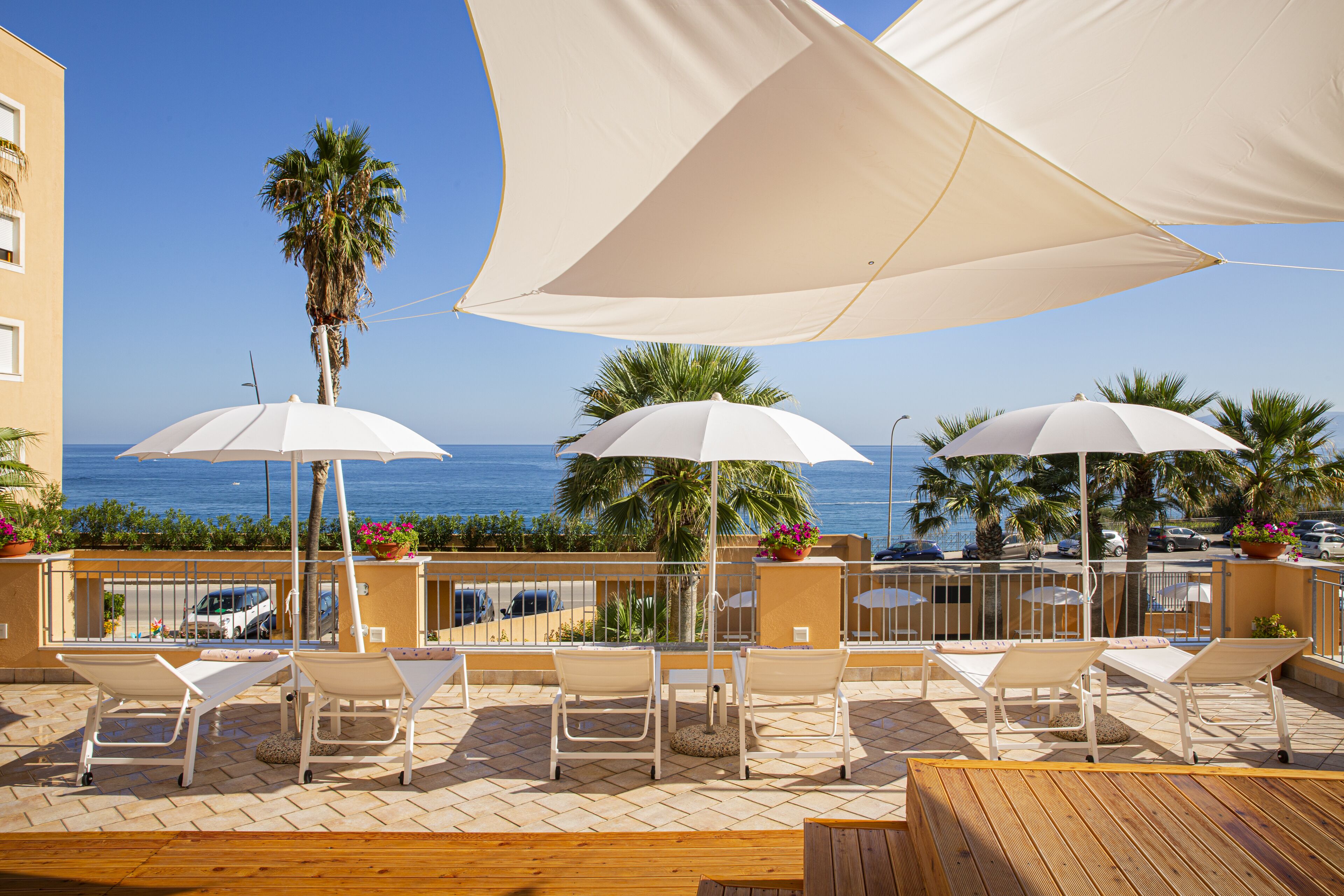 A terrace with white lounge chairs and large white umbrellas, overlooking a coastal area with palm trees and a clear blue sky.
