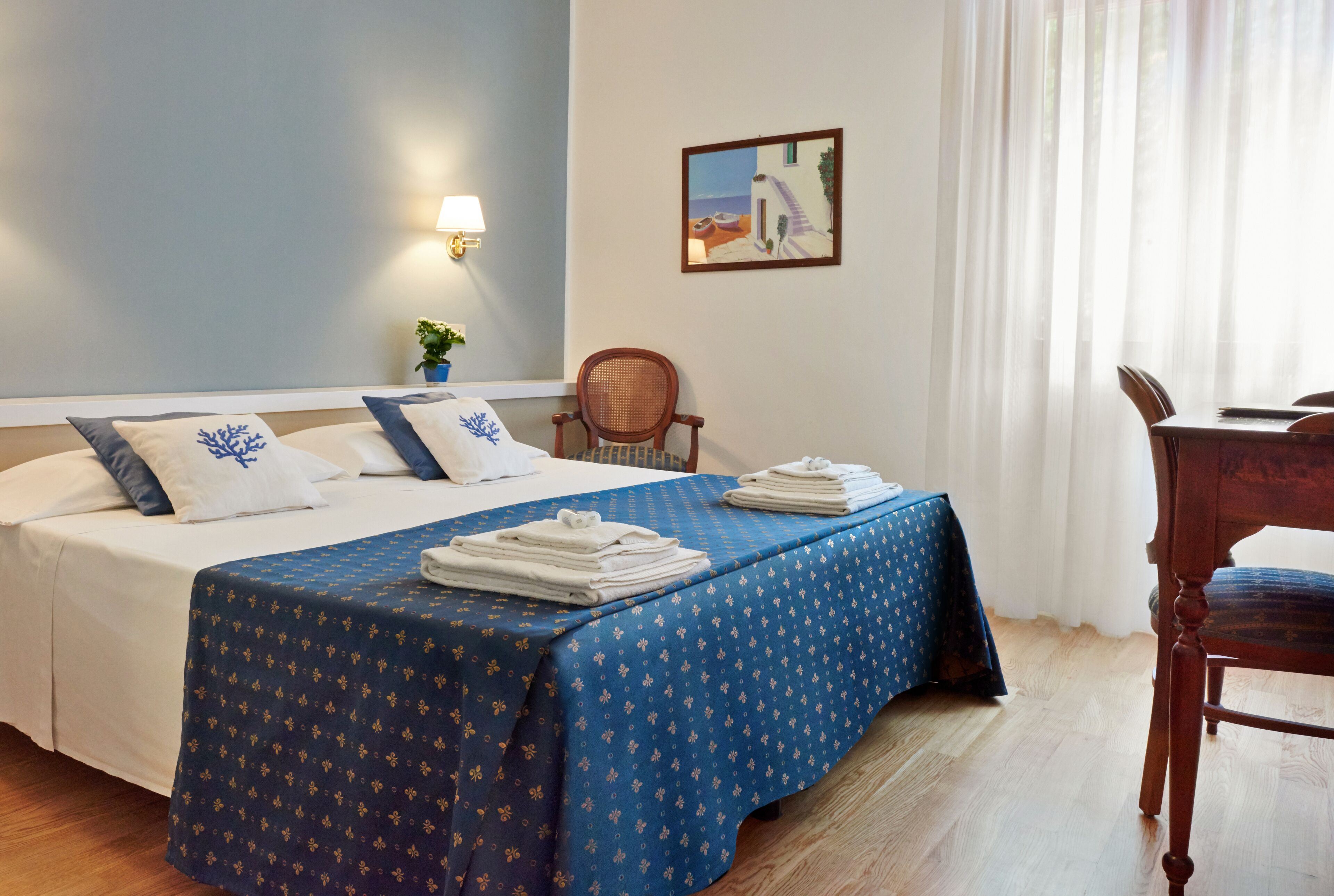 A hotel room with a bed, a chair, a table, a lamp, and a painting on the wall.