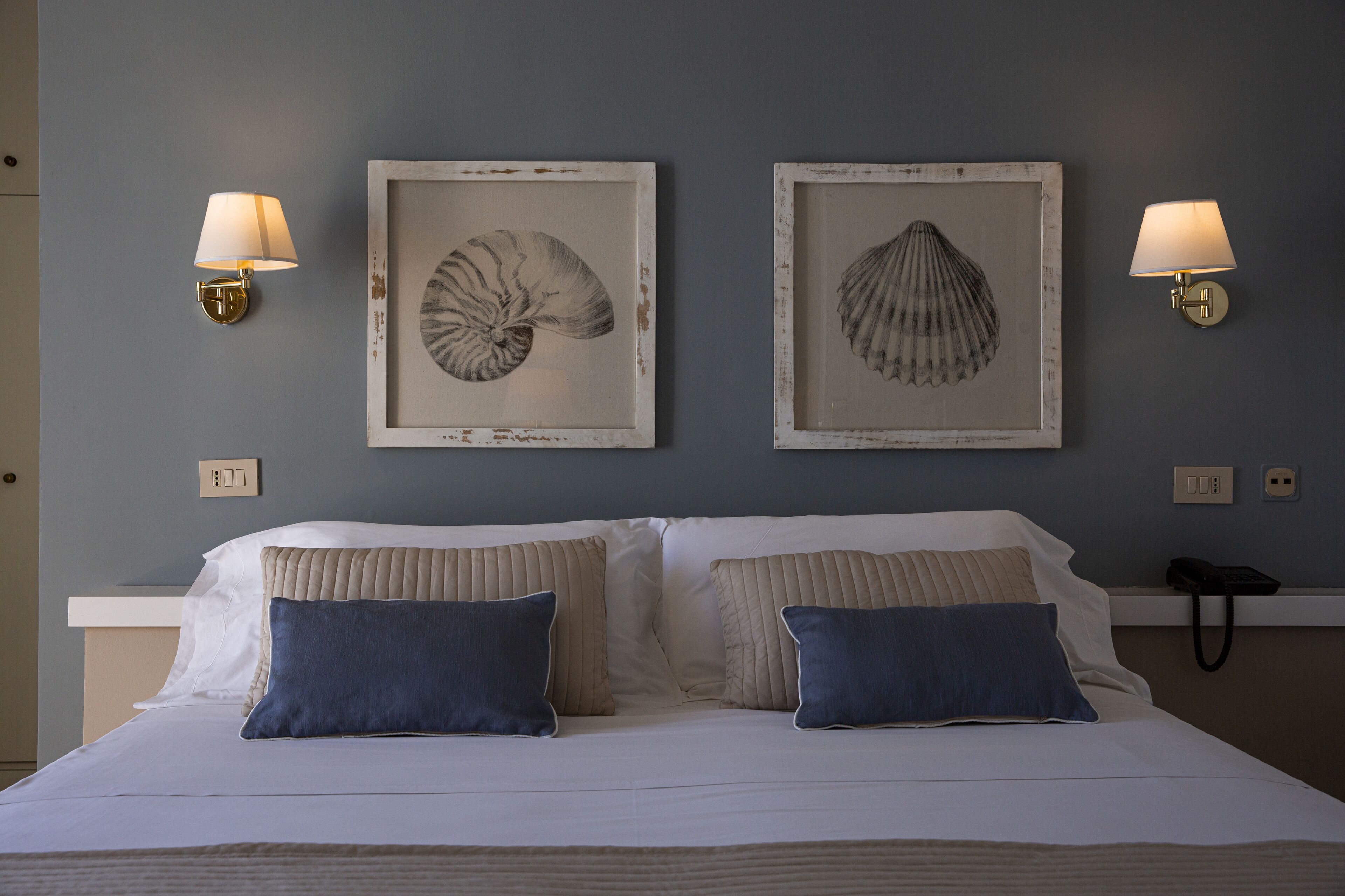 Uma cama com duas almofadas, uma parede com duas obras de arte com conchas emolduradas e duas luminárias fixadas na parede.