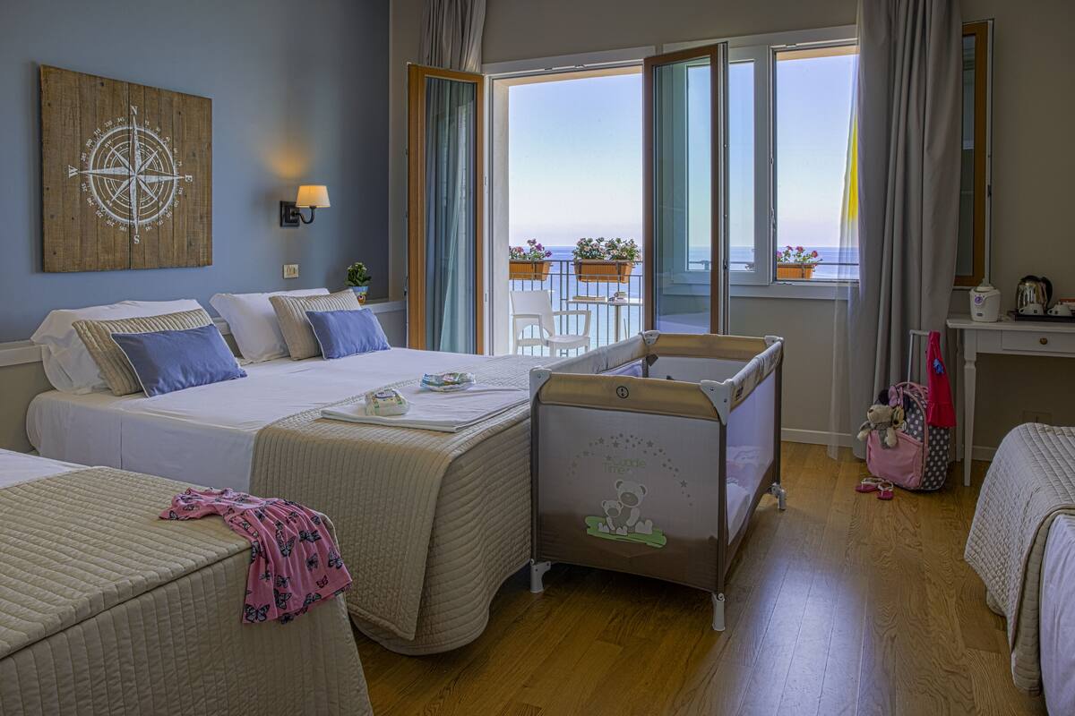 Habitación de hotel con dos camas, un balcón con vista al mar, un adorno de pared de madera y una mesita con una silla.