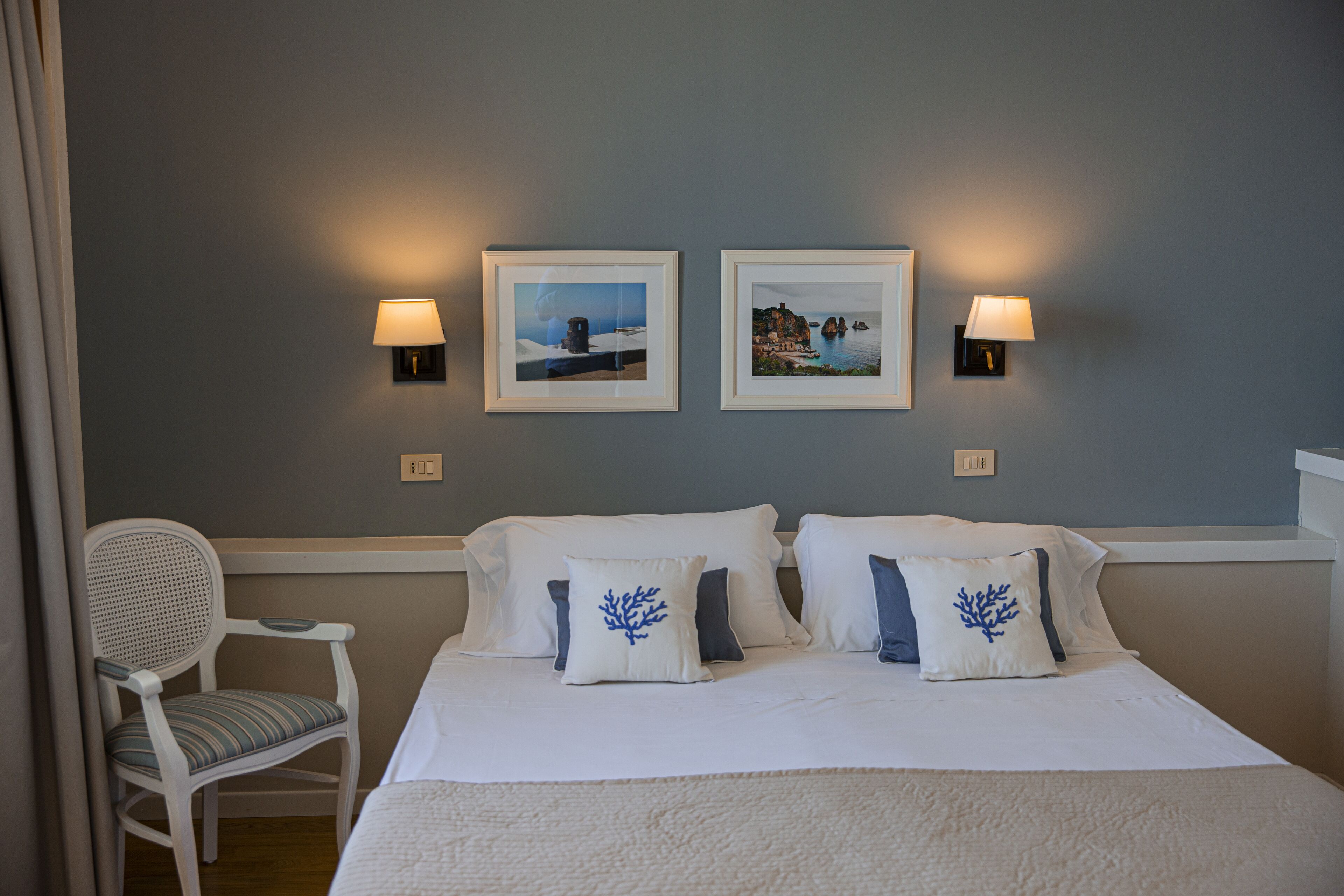 A hotel room with a bed, two wall-mounted lamps, two framed pictures, and a chair with a cushion.