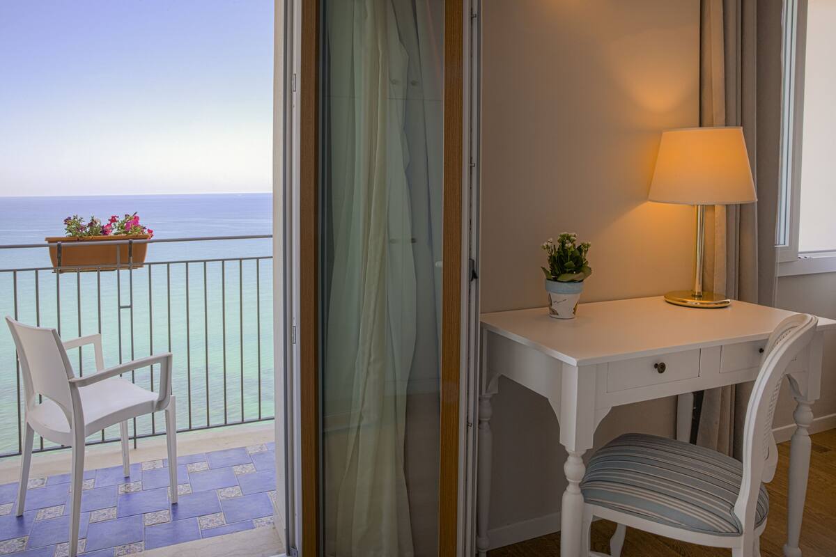 Un balcón con una silla y una planta en maceta, un escritorio blanco con una lámpara y vista al mar.