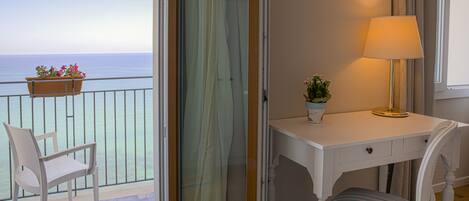 A balcony with a chair and a potted plant, a white desk with a lamp, and a view of the sea.