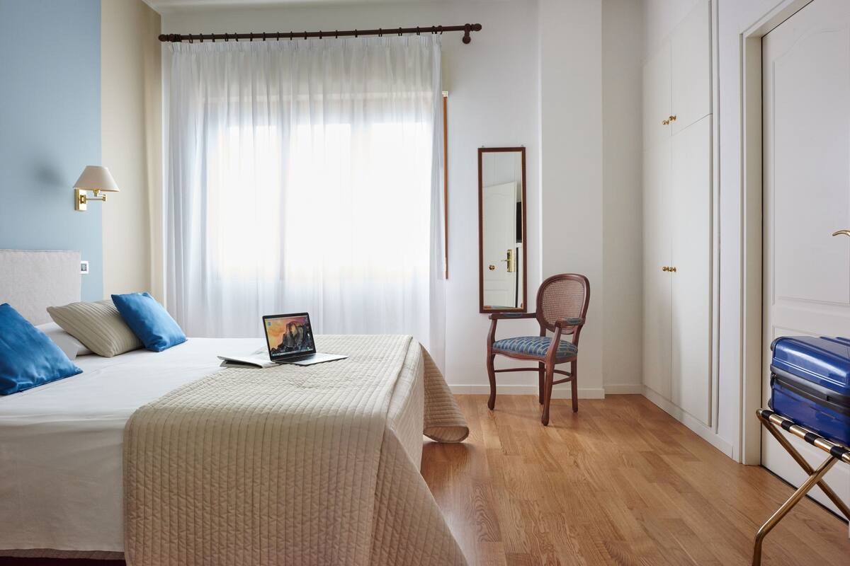 Una habitación de hotel con una cama, un escritorio con una computadora portátil, una silla y una maleta.