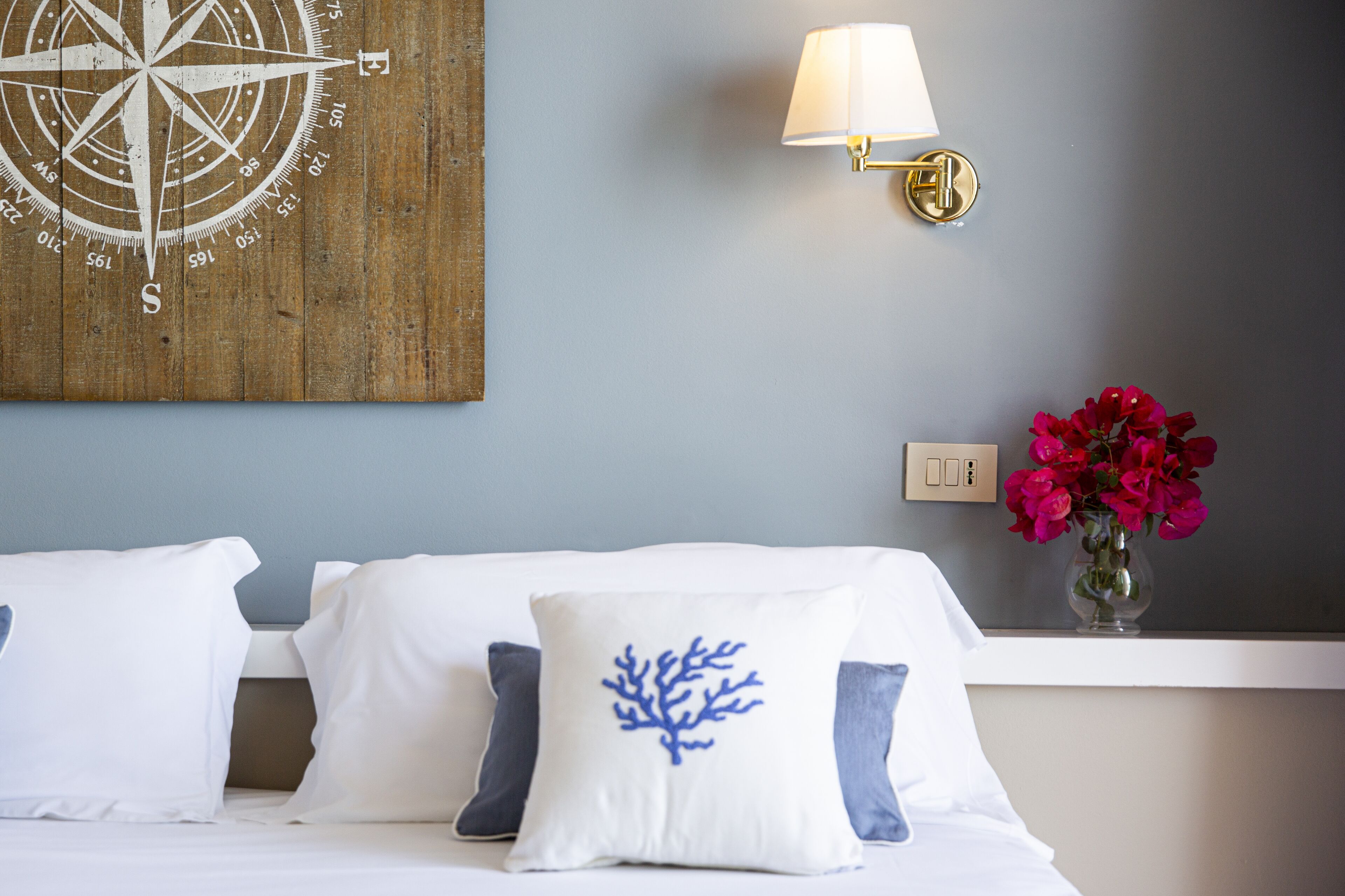 Una camera d'albergo con un letto, una bussola fissata al muro, una lampada da comodino, un vaso con fiori e una presa di corrente.