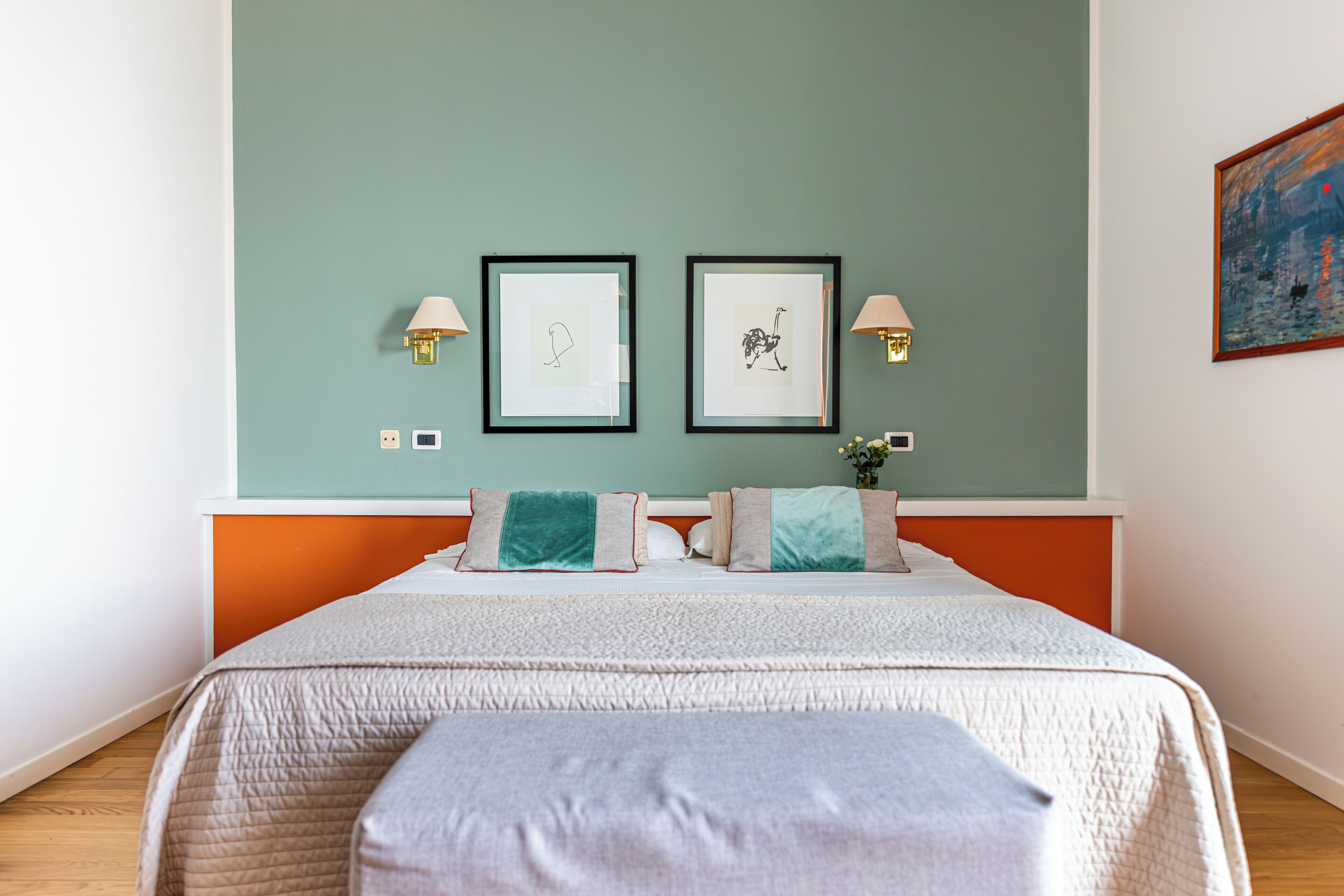 A neatly arranged bedroom with a bed, two framed artworks on the wall, and a nightstand with a lamp.