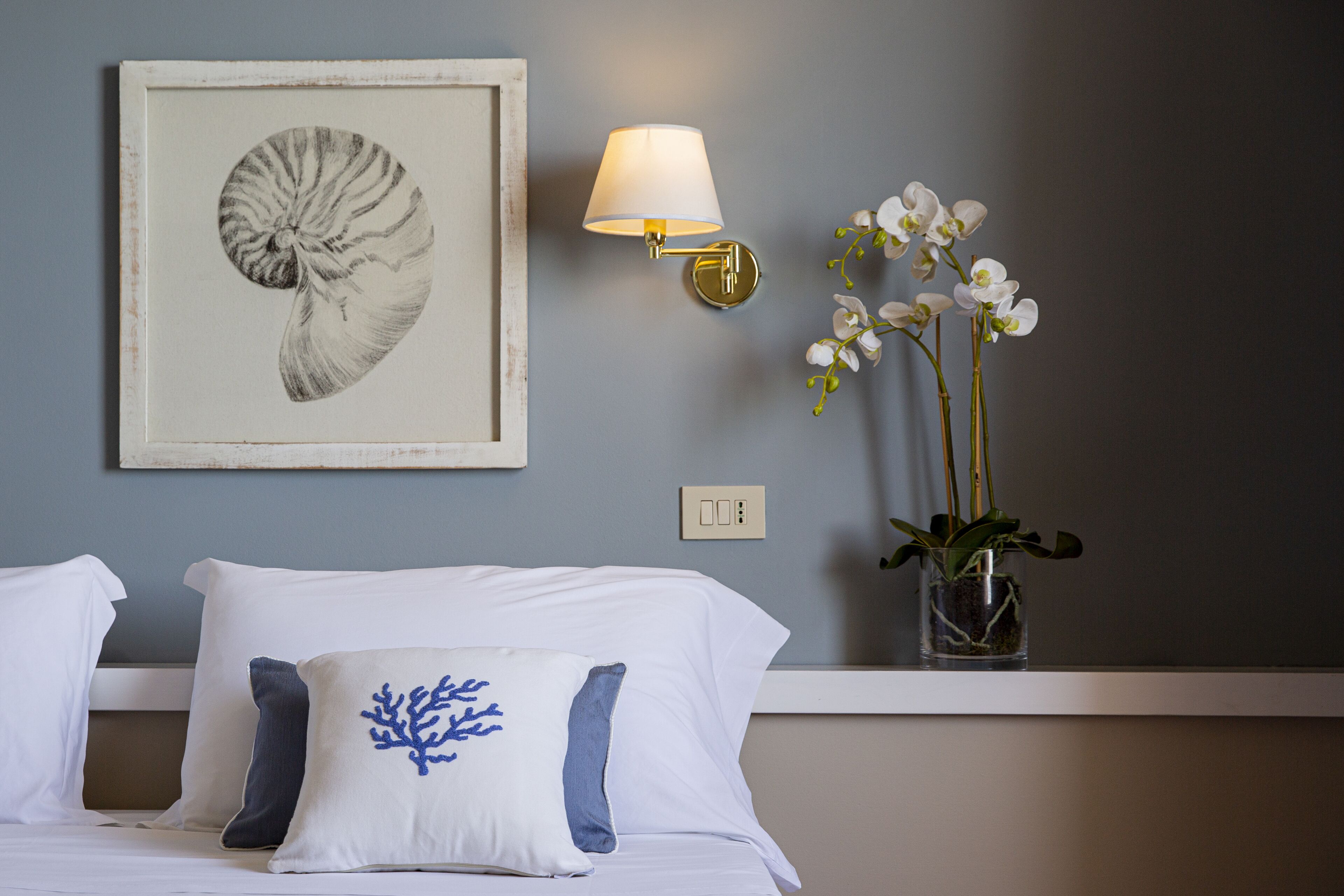 A hotel room with a bed, pillows, a framed artwork, a wall-mounted lamp, and a vase with flowers.
