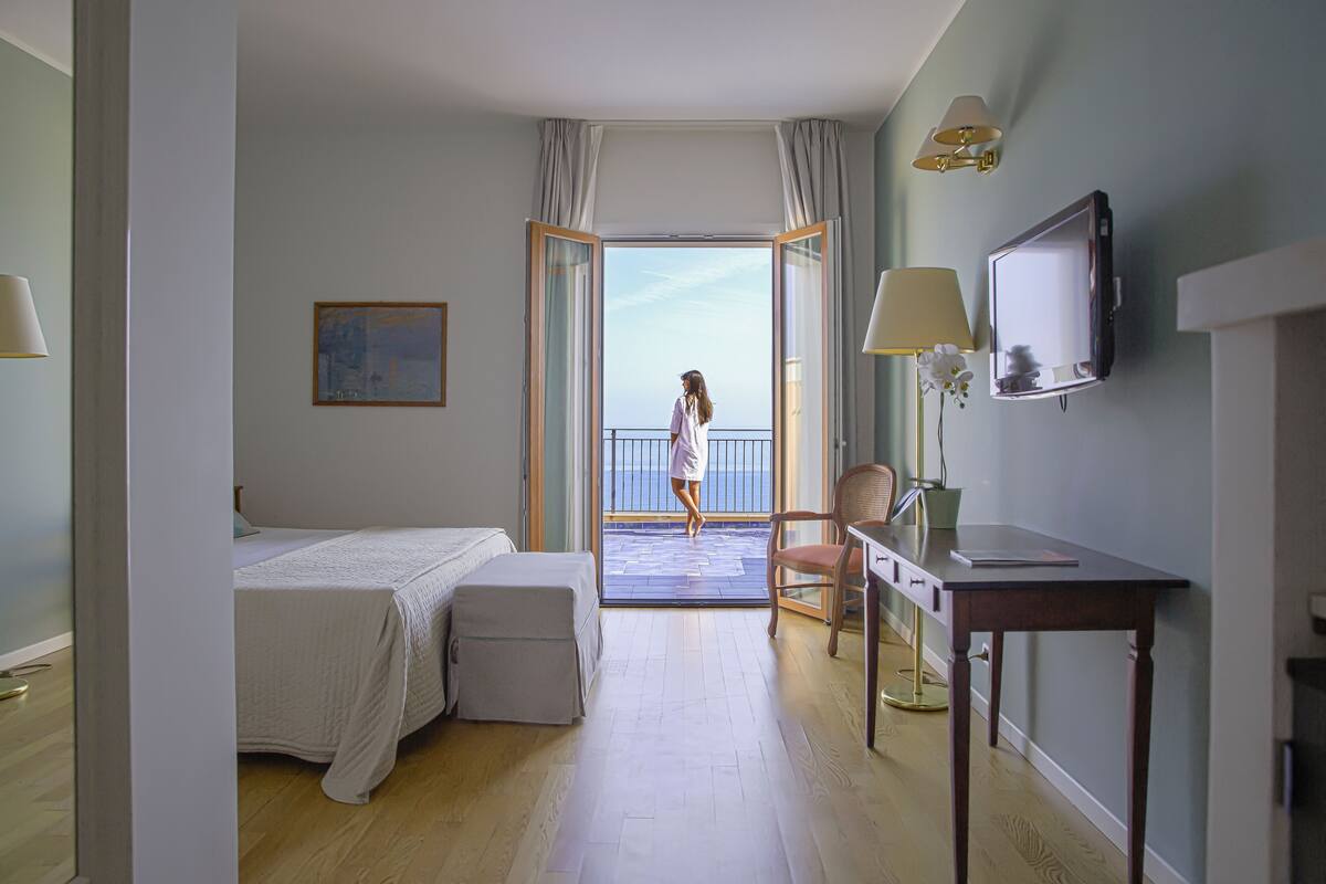 Habitación de hotel con cama, escritorio, silla, televisión y balcón con vistas.
