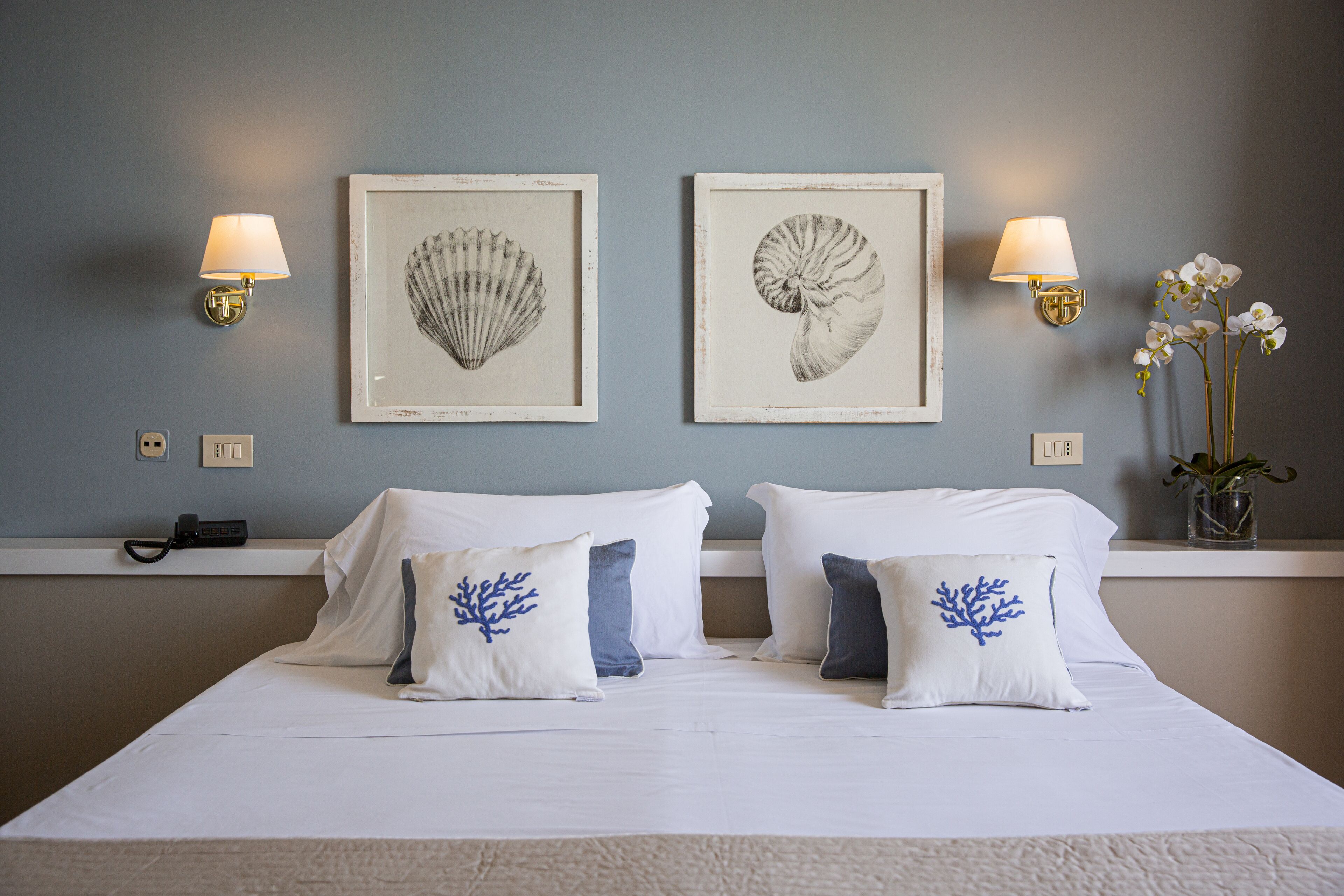A hotel room with a bed, two wall-mounted lamps, and two framed artwork depicting shells.