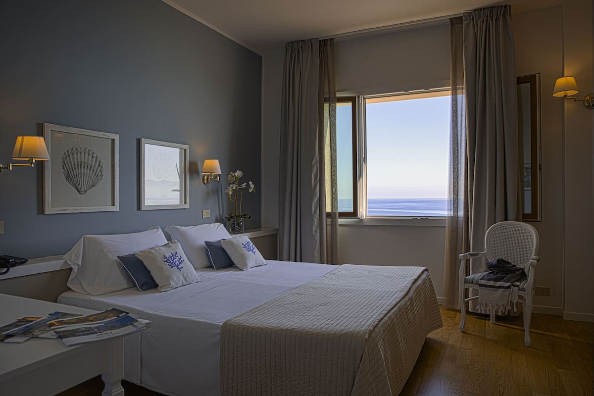 Una habitación de hotel con una cama grande, una silla, un ventanal con vistas y lámparas de pared.