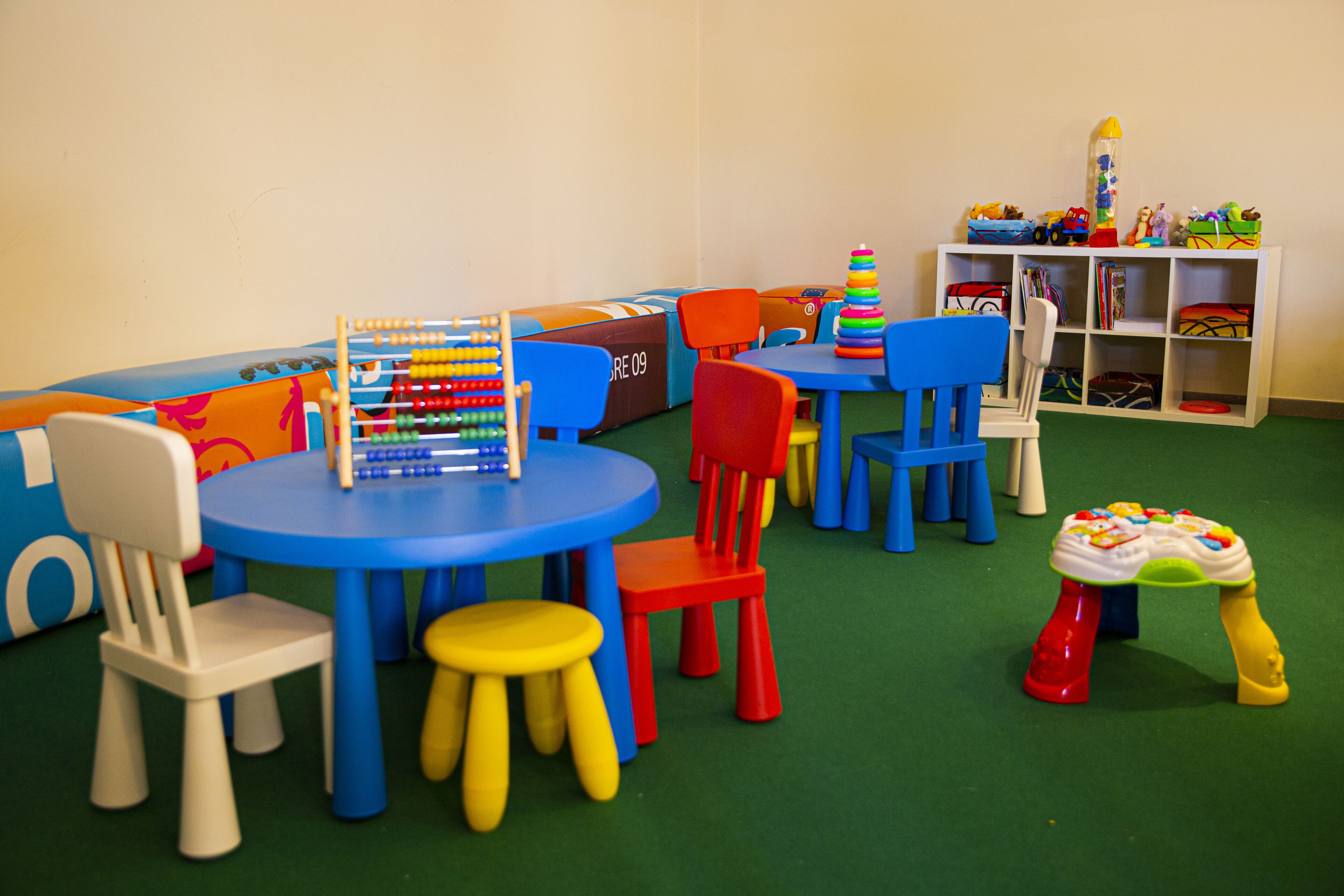 Een speelruimte voor kinderen, met een blauwe tafel, rode en gele krukken en een witte plank met speelgoed.