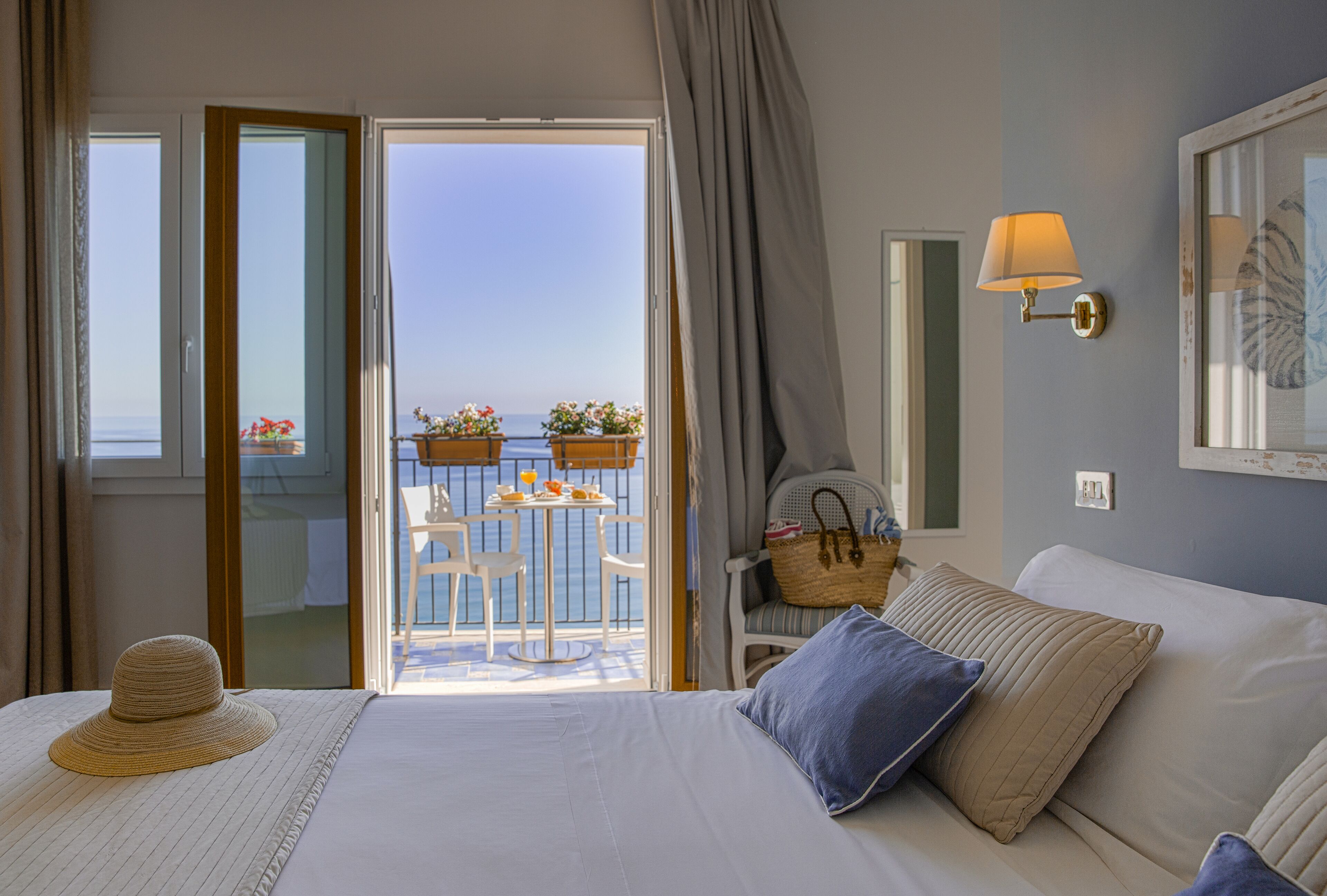 A hotel room with a bed, a view of the sea, a balcony with a table and chairs, and a wall-mounted lamp.