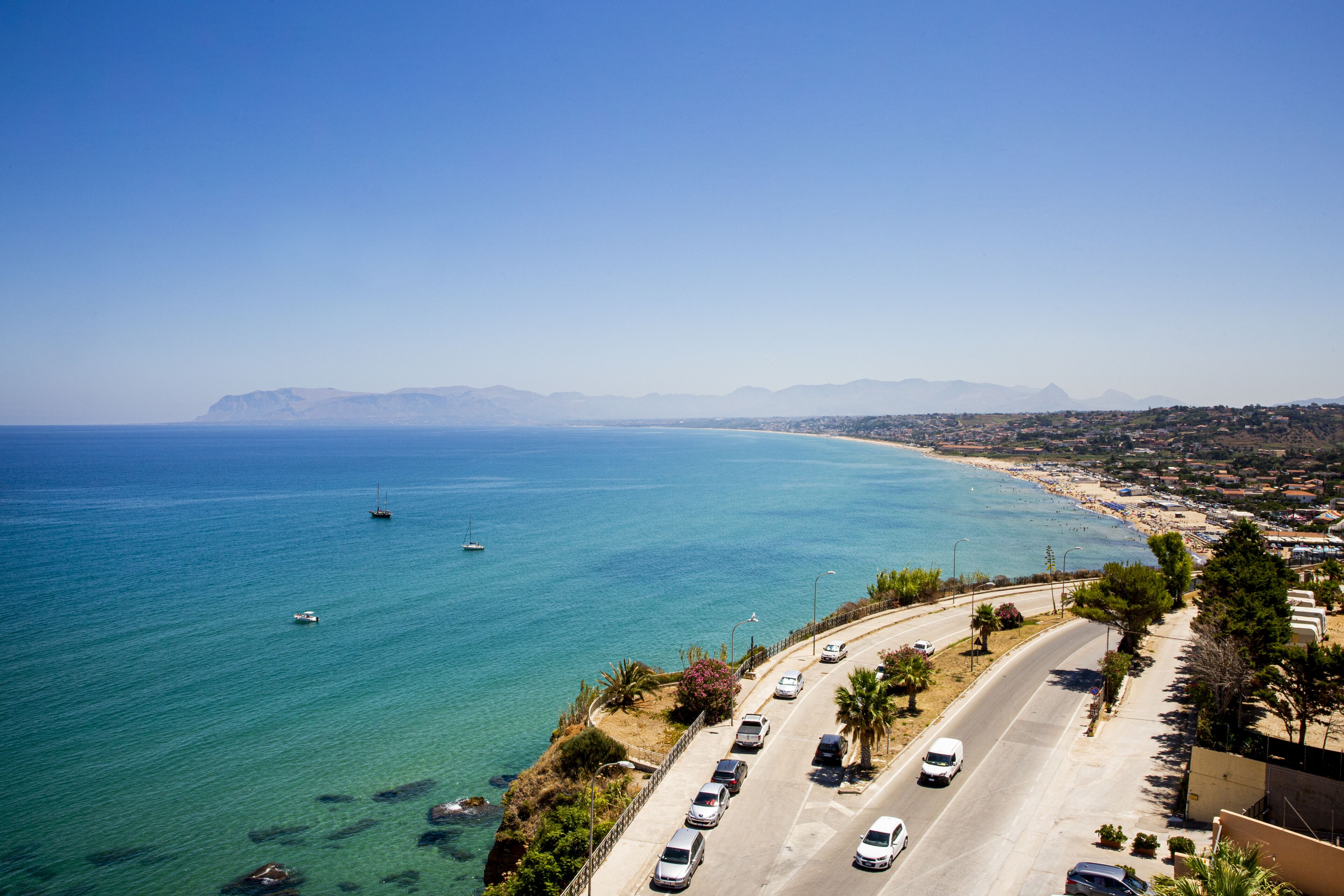 A coastal road with parked cars, a clear blue sea, and distant mountains.