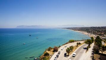 A coastal road with parked cars, a clear blue sea, and distant mountains.