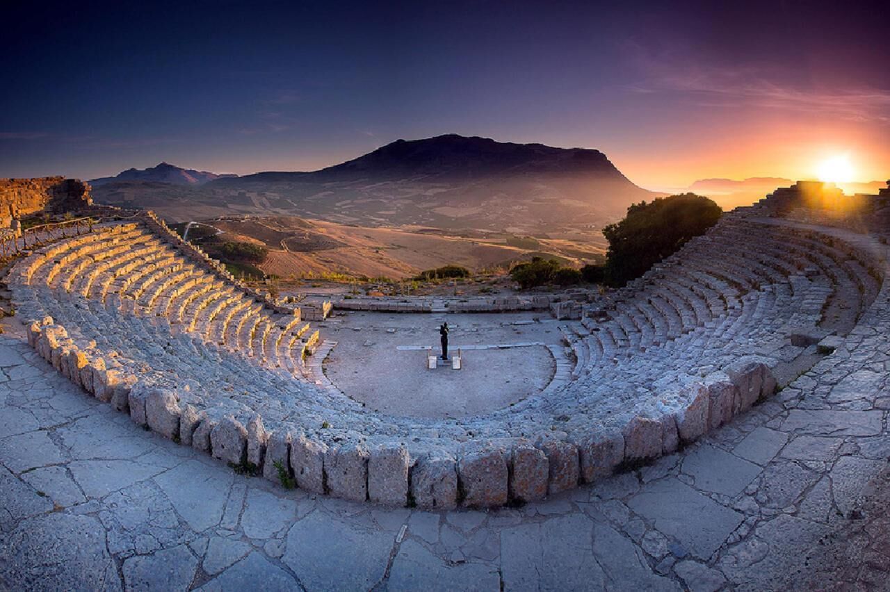Un antiguo anfiteatro con asientos de piedra y un escenario central, rodeado por un paisaje montañoso.