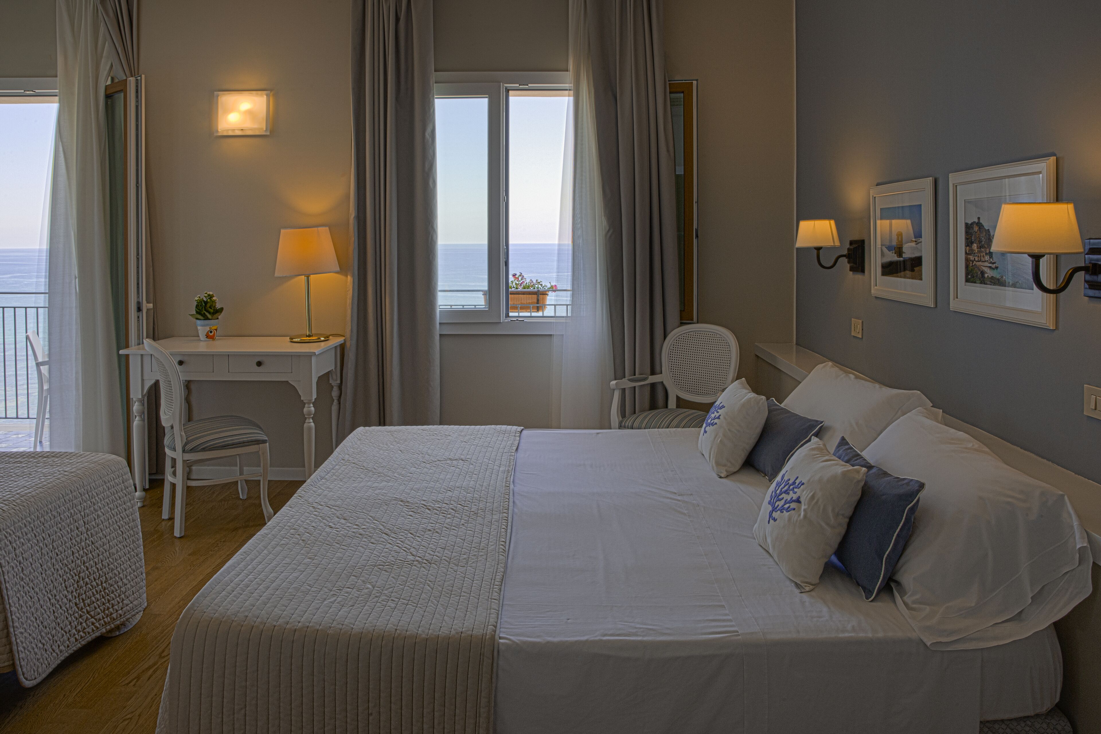 A hotel room with a large bed, a desk, a chair, and a view of the sea through the window.