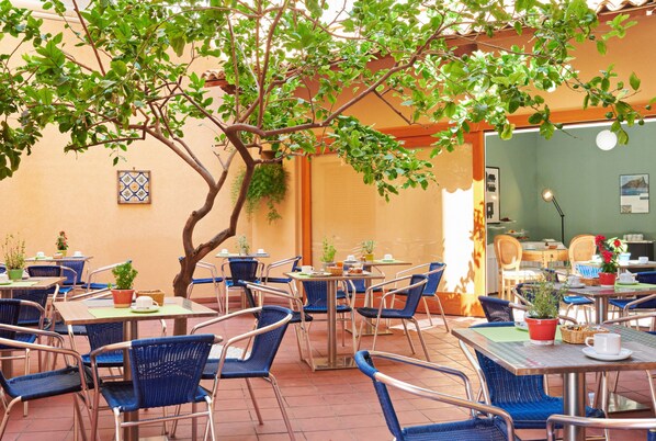 Un café al aire libre con mesas y sillas, rodeado de follaje y un piso de baldosas.