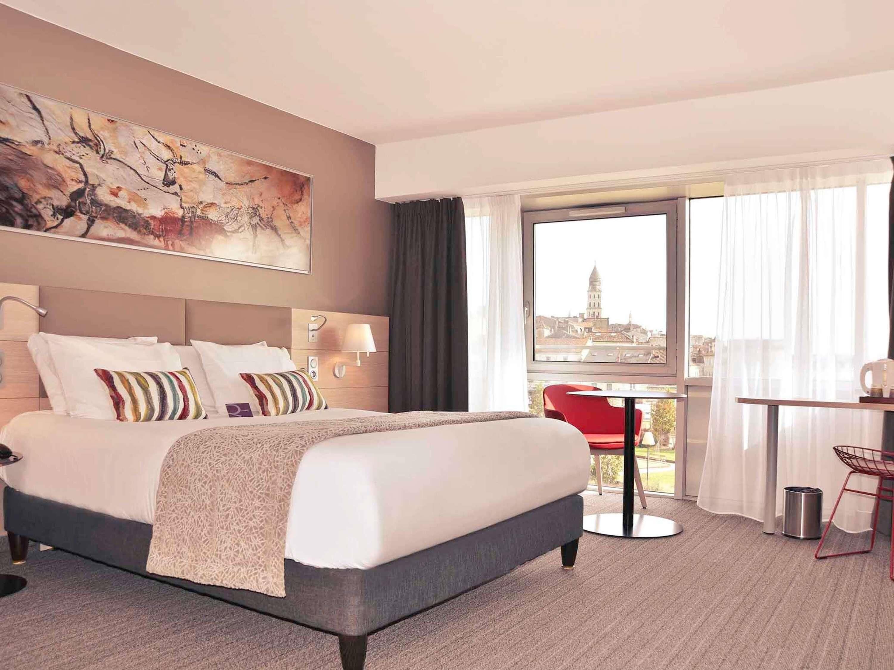 Habitación de hotel con cama, escritorio, silla, vista a la ciudad y un cuadro en la pared.