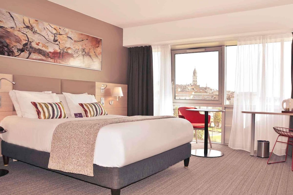 Habitación de hotel con cama, escritorio, silla, vista a la ciudad y un cuadro en la pared.