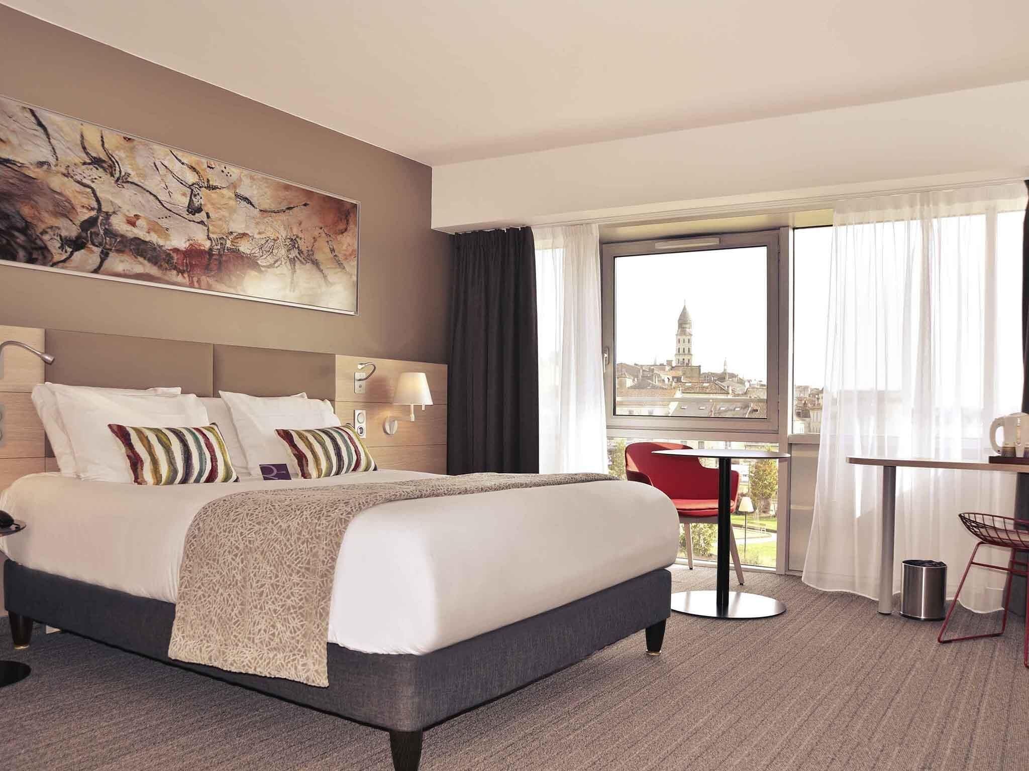 A hotel room with a bed, a desk, a chair, a view of a cityscape, and a painting on the wall.