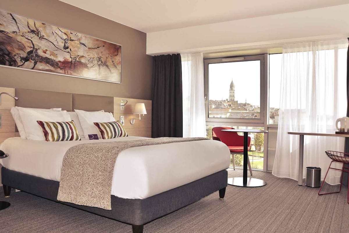 A hotel room with a bed, a desk, a chair, a view of a cityscape, and a painting on the wall.