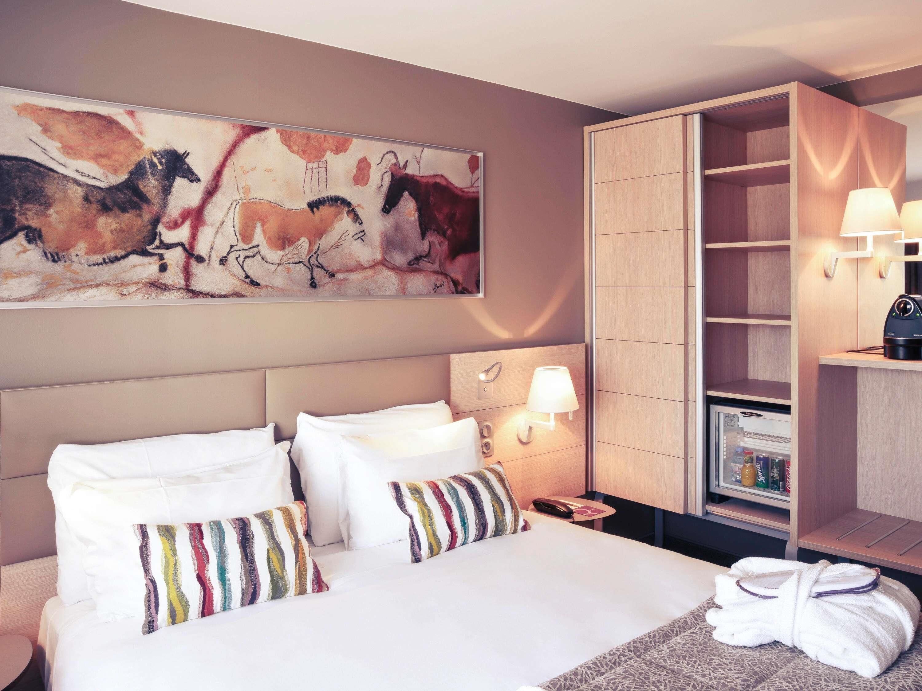 A hotel room with a bed, pillows, a nightstand, a lamp, a robe, a coat rack, and a painting on the wall.