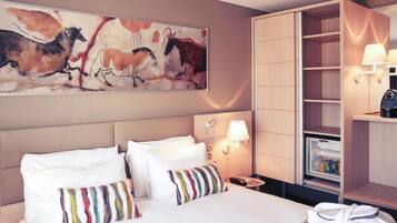 A hotel room with a bed, pillows, a nightstand, a lamp, a robe, a coat rack, and a painting on the wall.